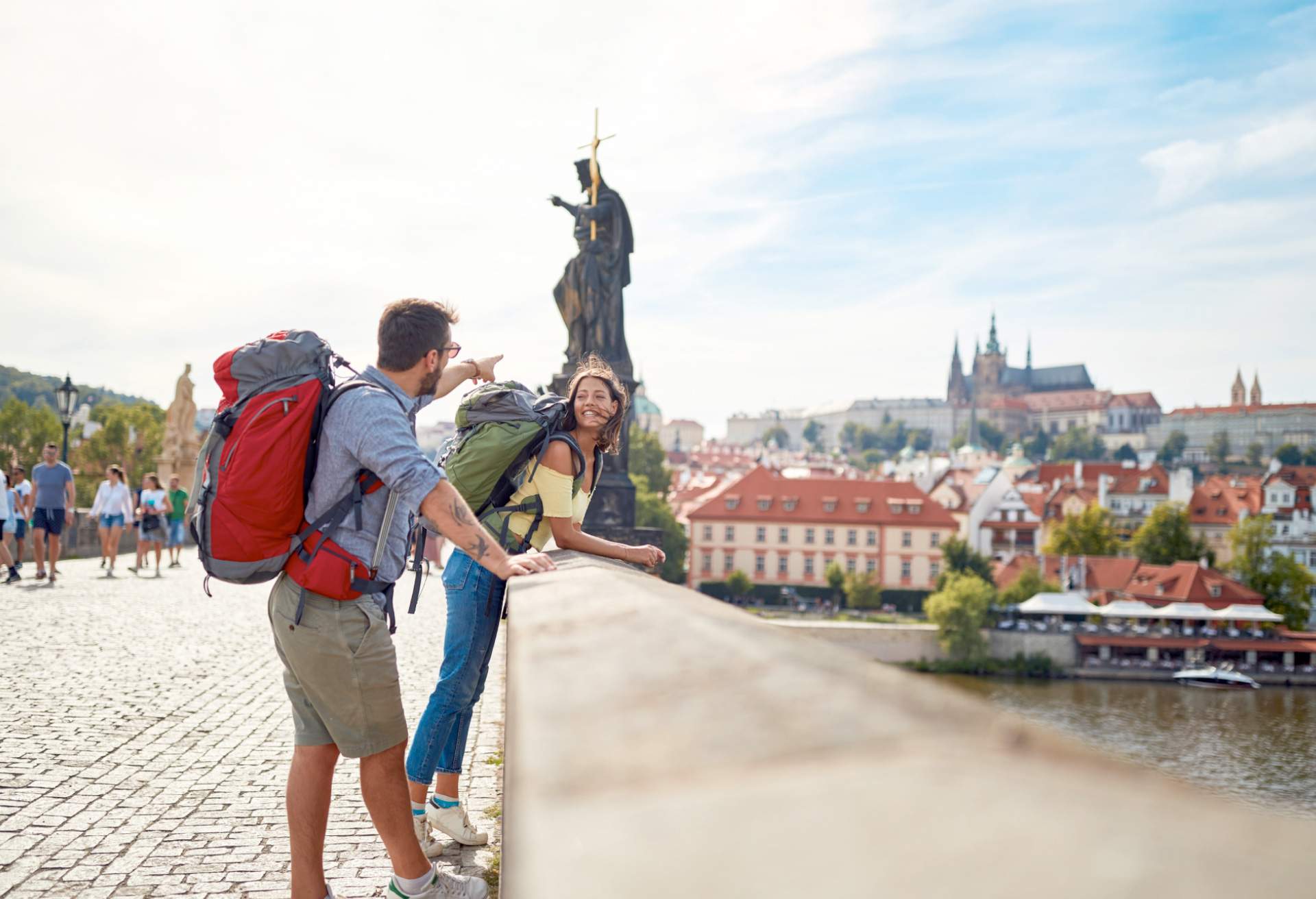 A young man and woman lean over a bridge in Europe. The man points at the view in the distance while the woman looks at him, smiling.