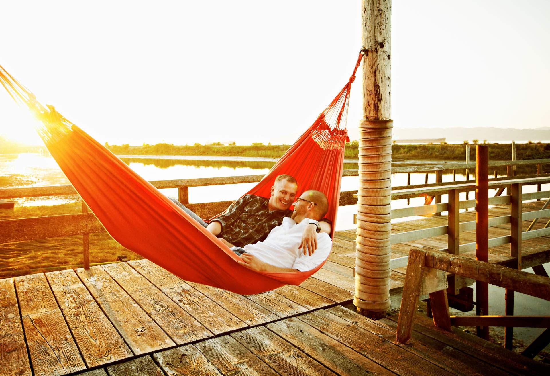Two men lay together in a hammock, smiling, as the sun sets behind them