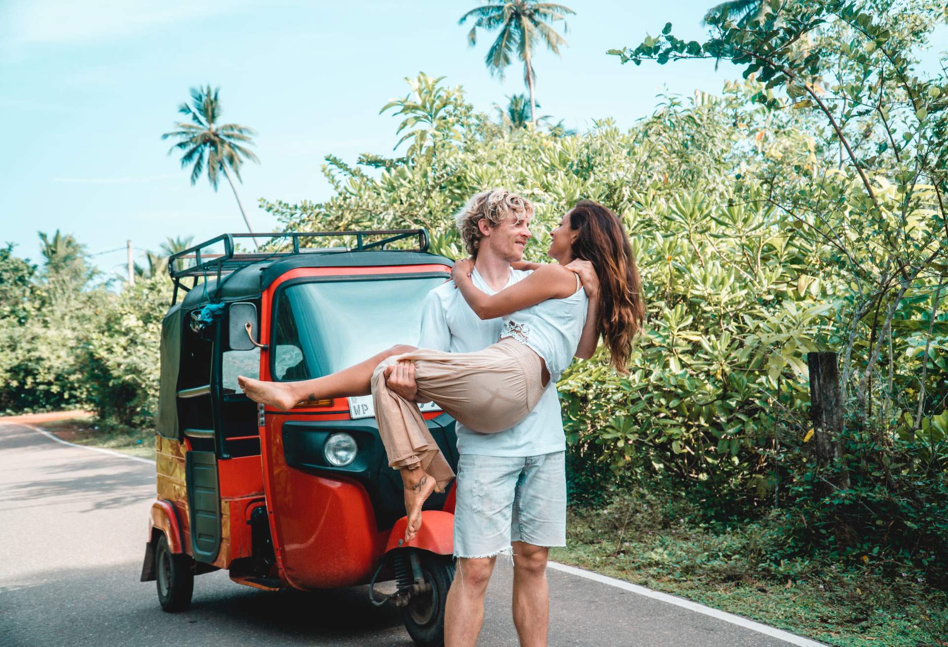 A man and woman couple stand in front of a red tuk-tuk with palm trees lining the road. The pan is cradling the woman in his arms.