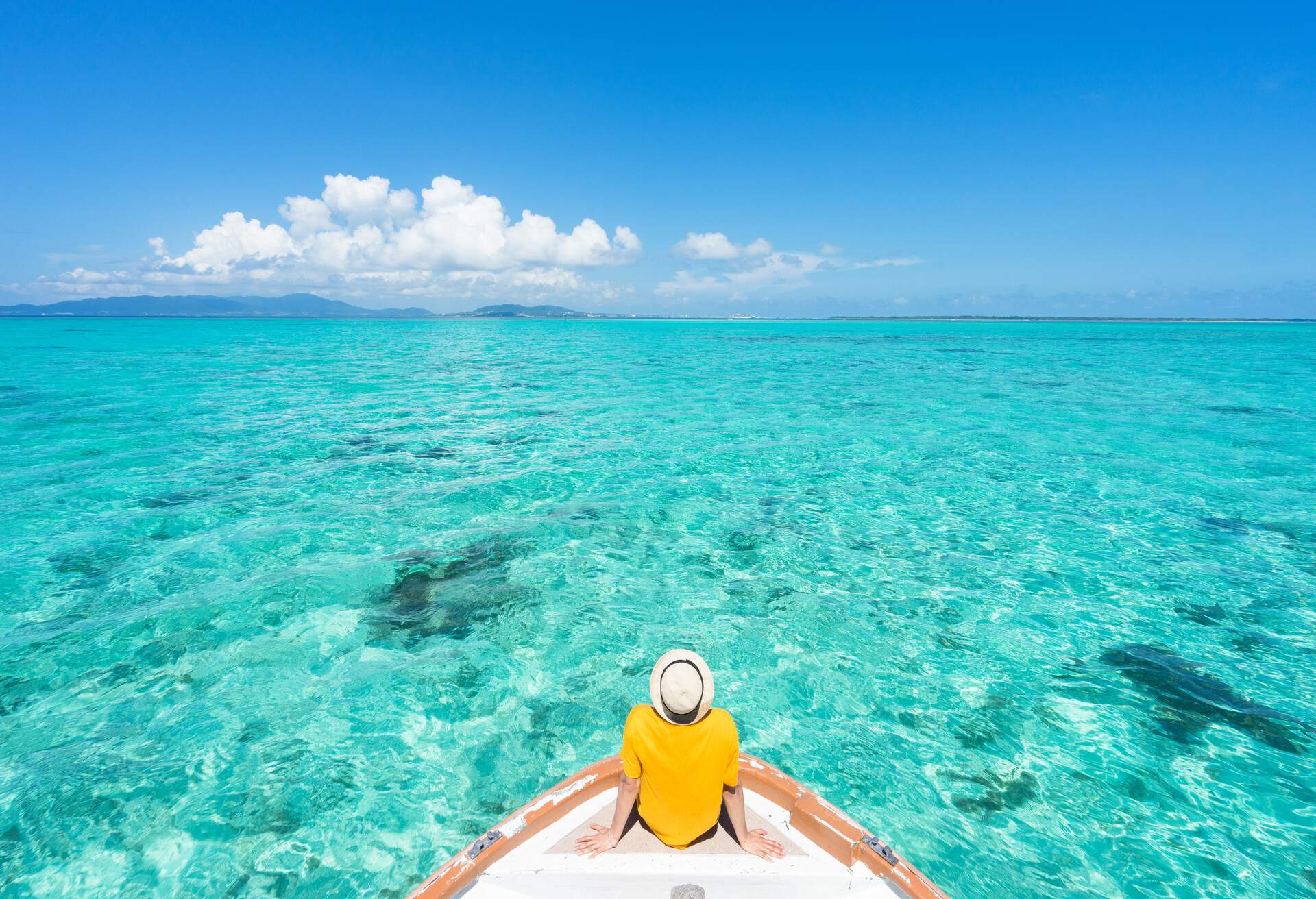 A man in a yellow shirt and a hat sitting on the prow of a boat, gazing out into the aquamarine waters.