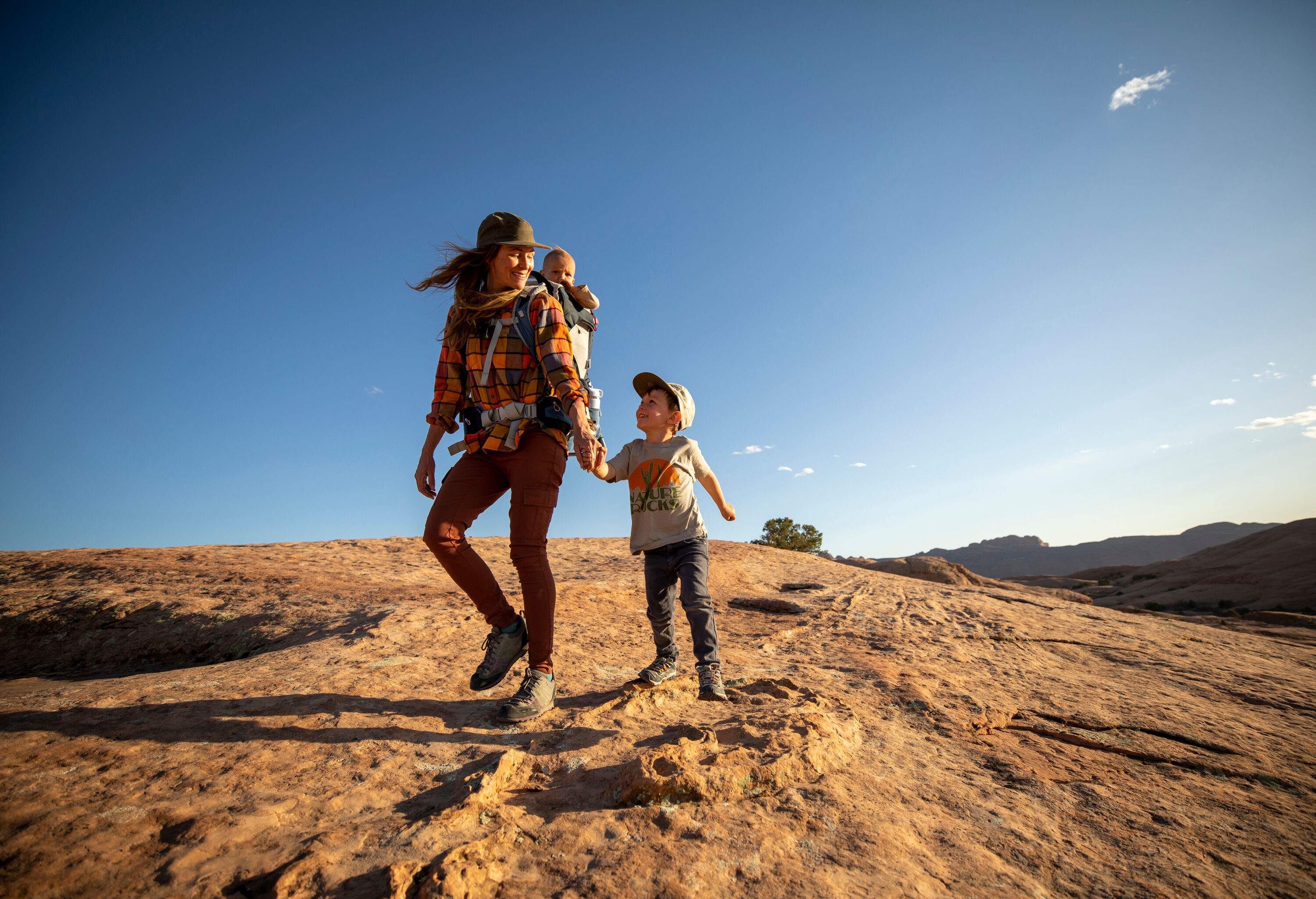 A woman hiking on a sunny day with a baby on her back and a little boy holding her hand.