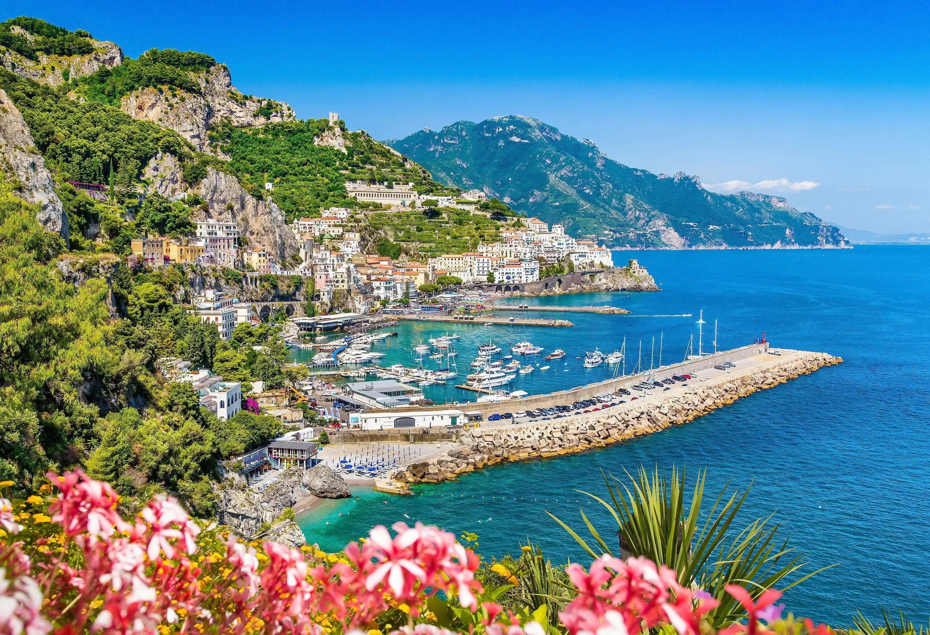 A vista of the Amalfi Coast surrounded by beautiful greenery, with boats docked in the port and a breakwater in the background, and steep slopes in the front.