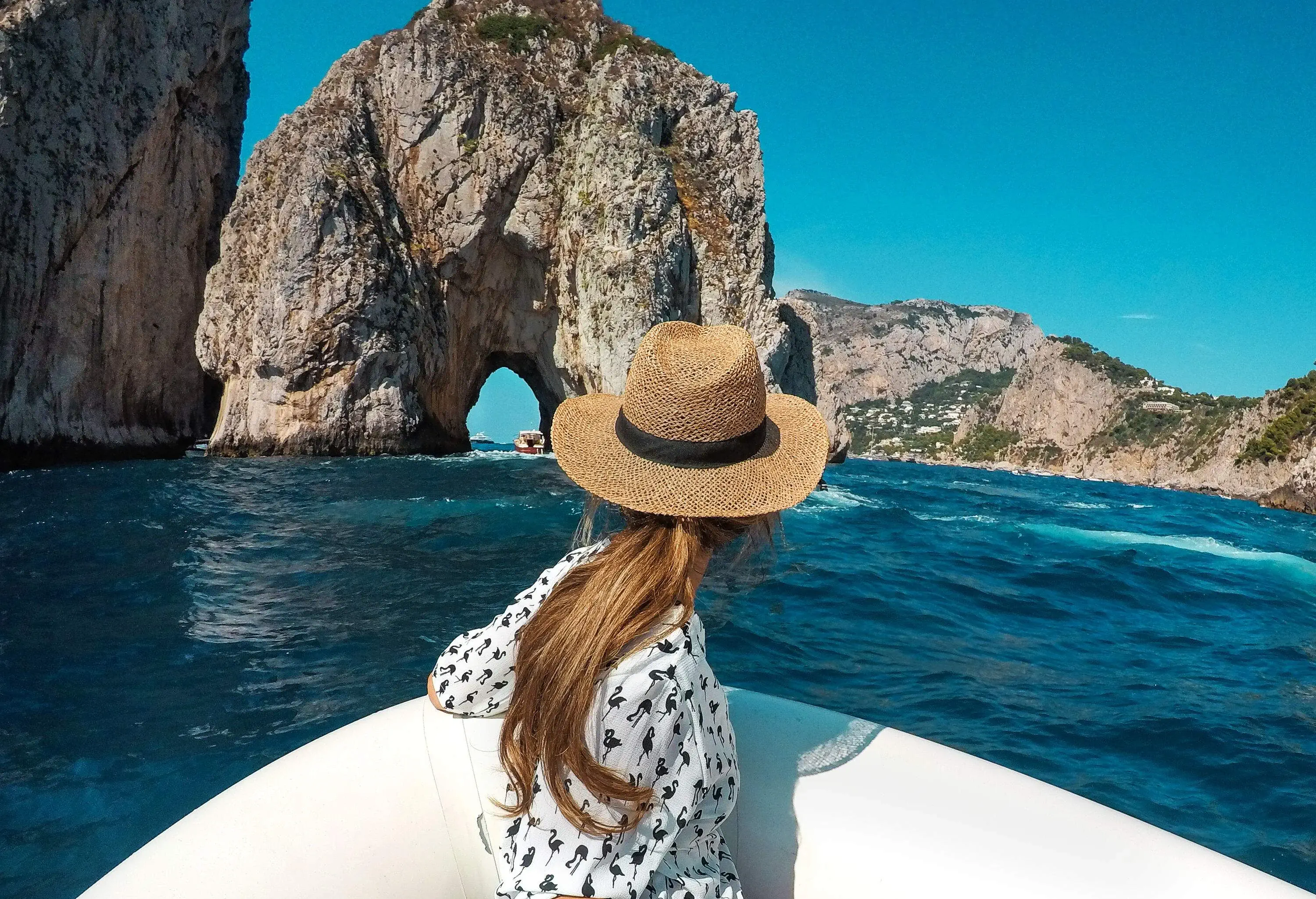 A brunette woman in a printed dress and straw hat rides a white boat and glances back at a rock formation with an arch entrance.