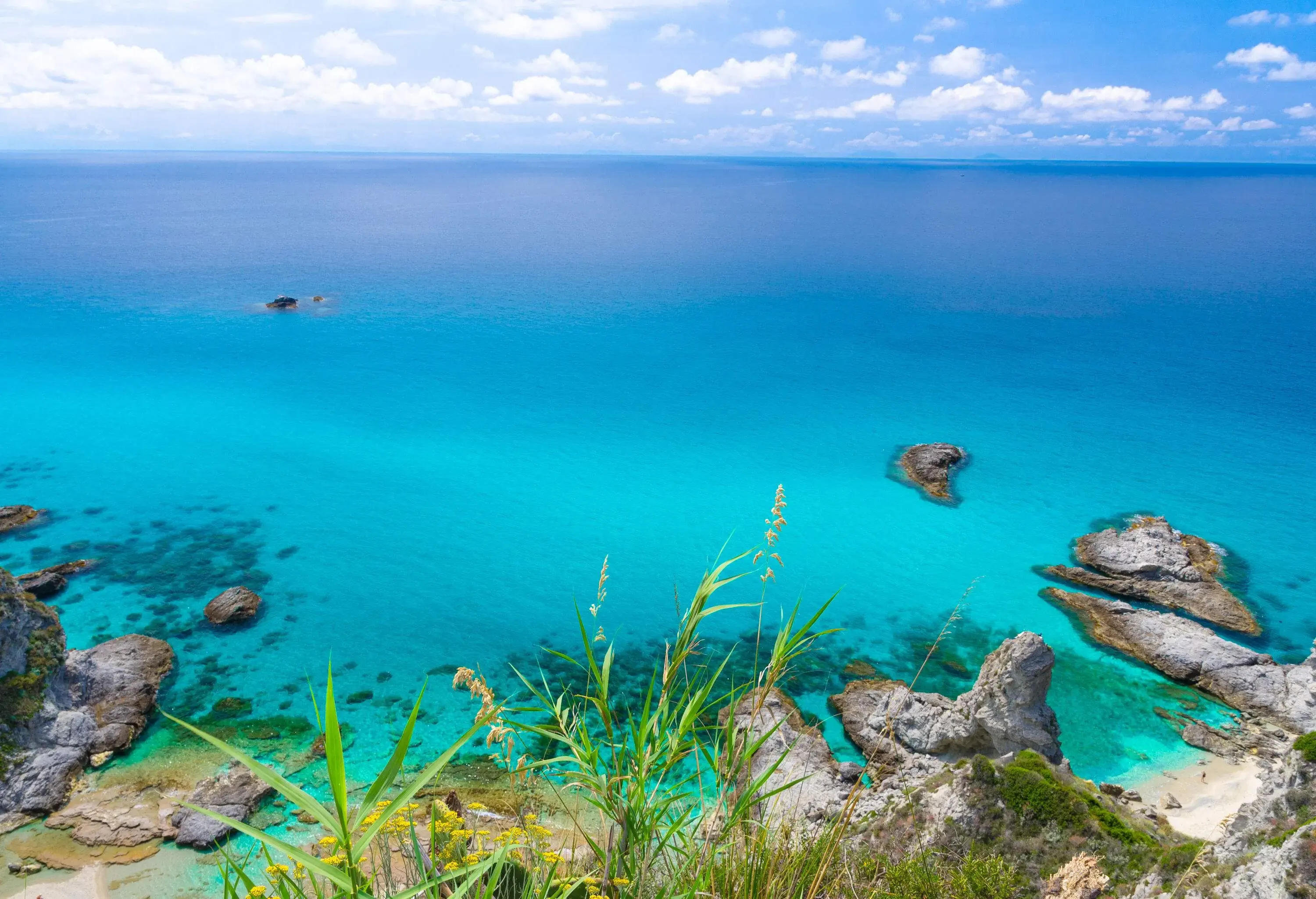 A rocky coastline with views of a vast azure ocean as seen from a grassy cliff.