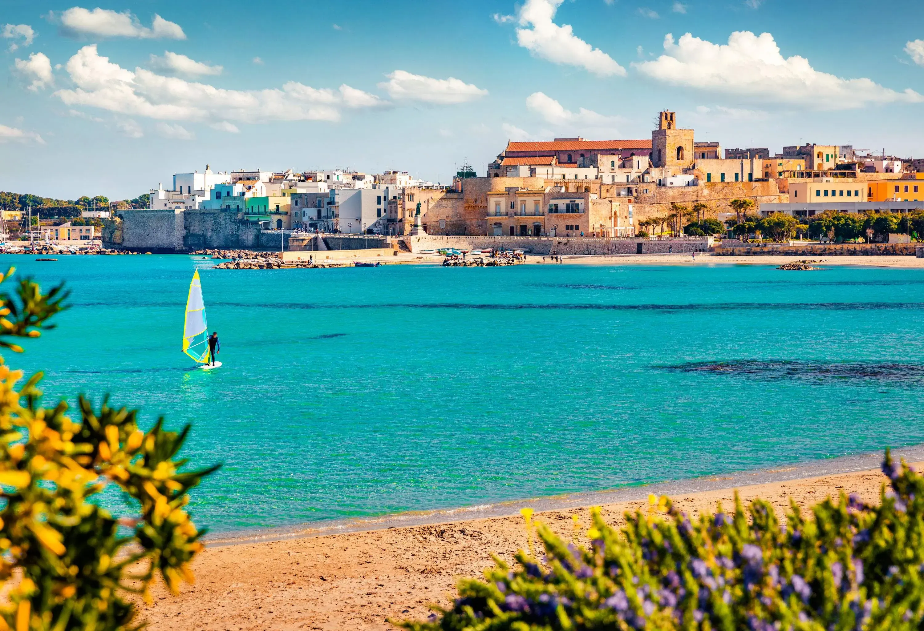 A man stands on a windsurfer on a tranquil turquoise beach with compact houses and buildings on the coast in the background.