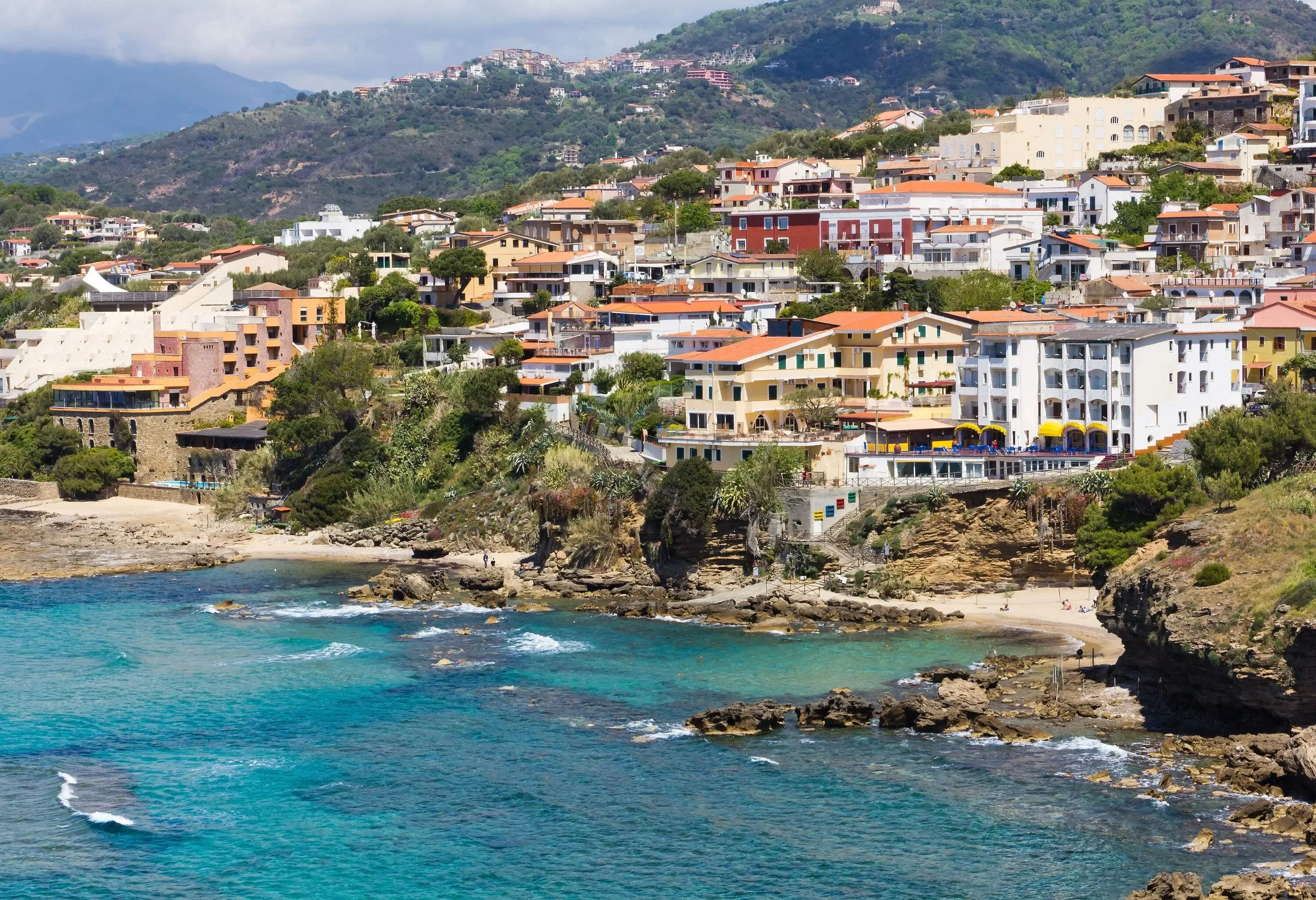 A coastal town situated on a slope with a lush green hill visible in the background.