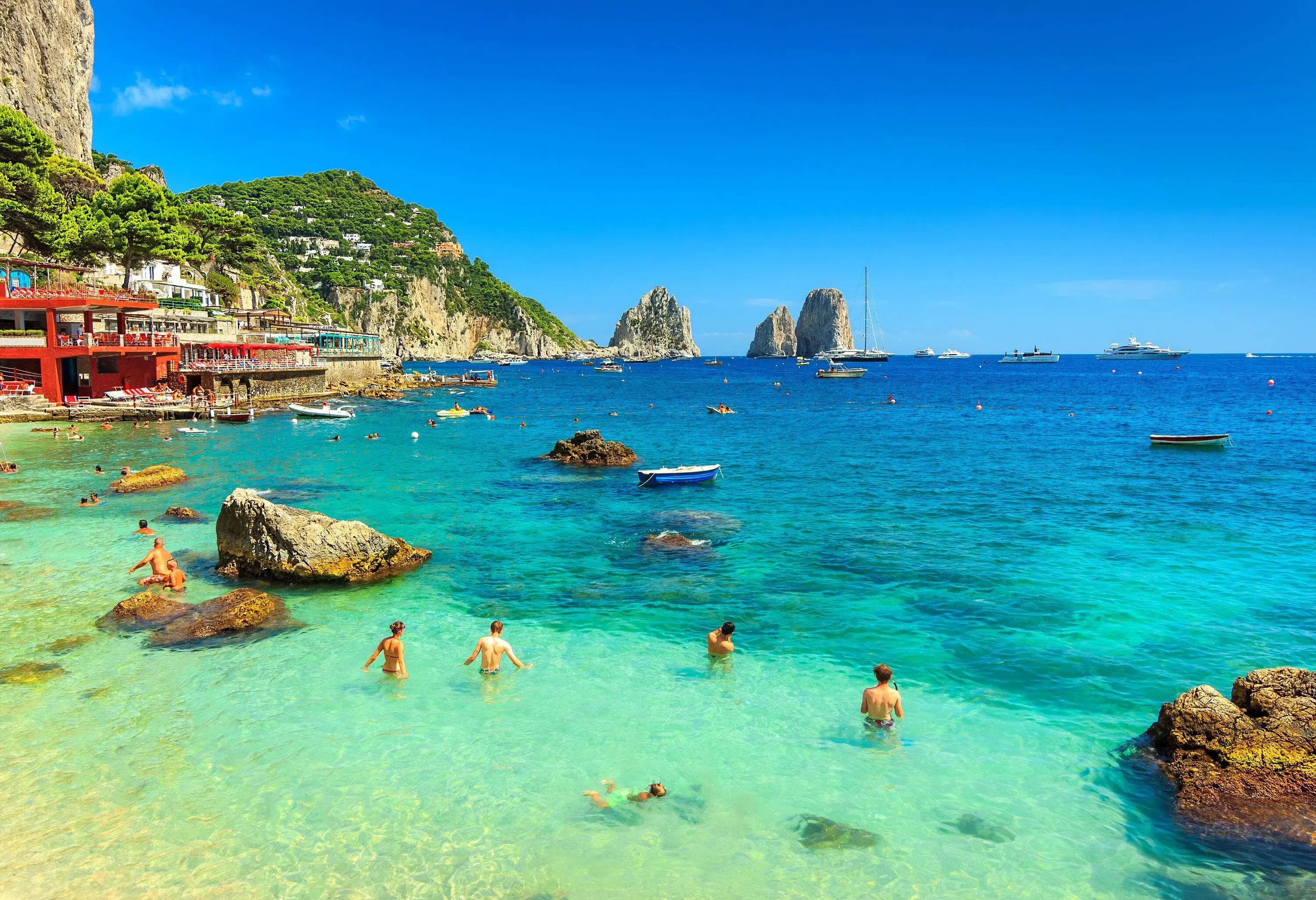People swimming in a crystal-clear sea littered with large boulders.