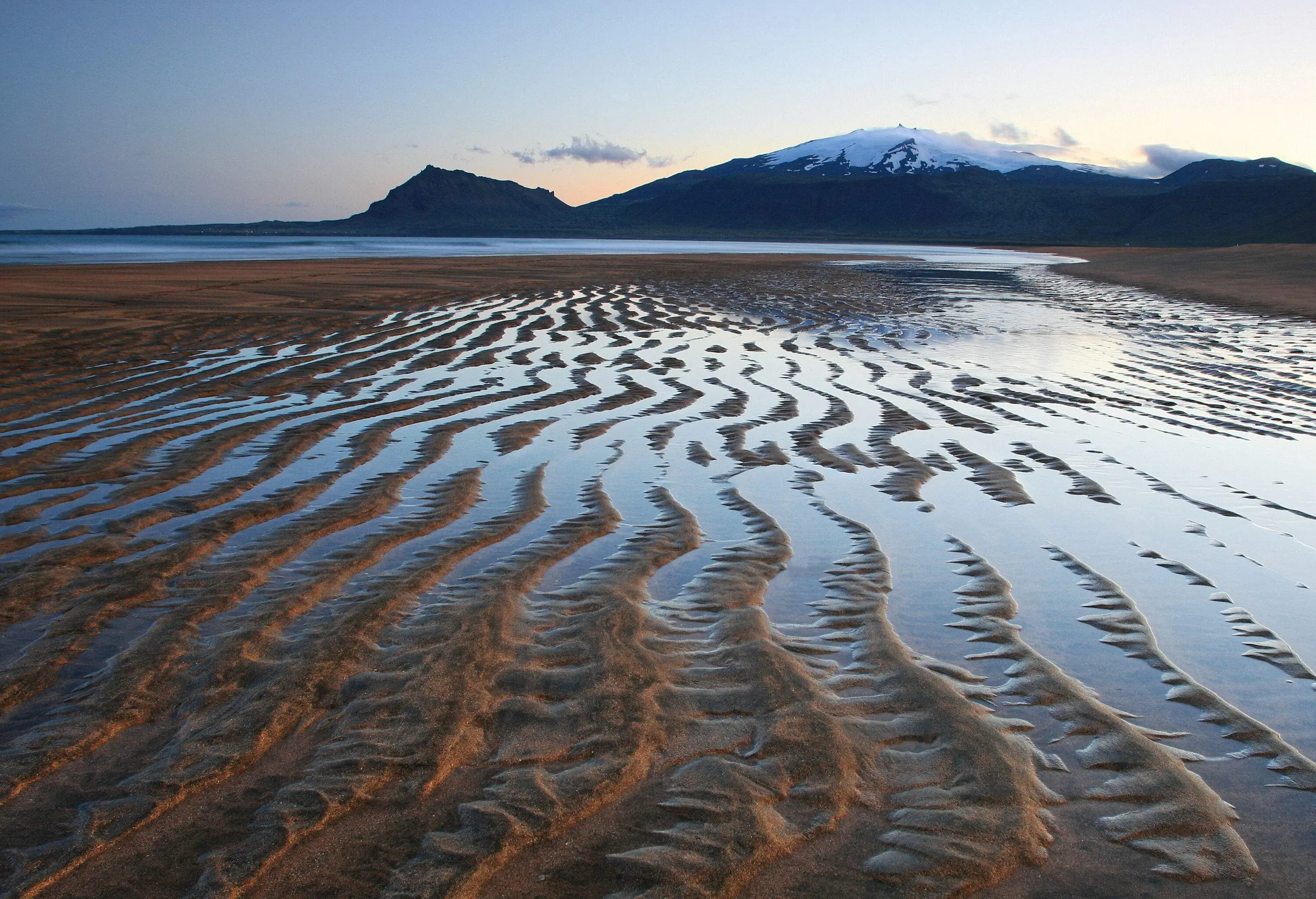 Tidal flats with puddles of water on the beach and a snow-capped mountain in the background.