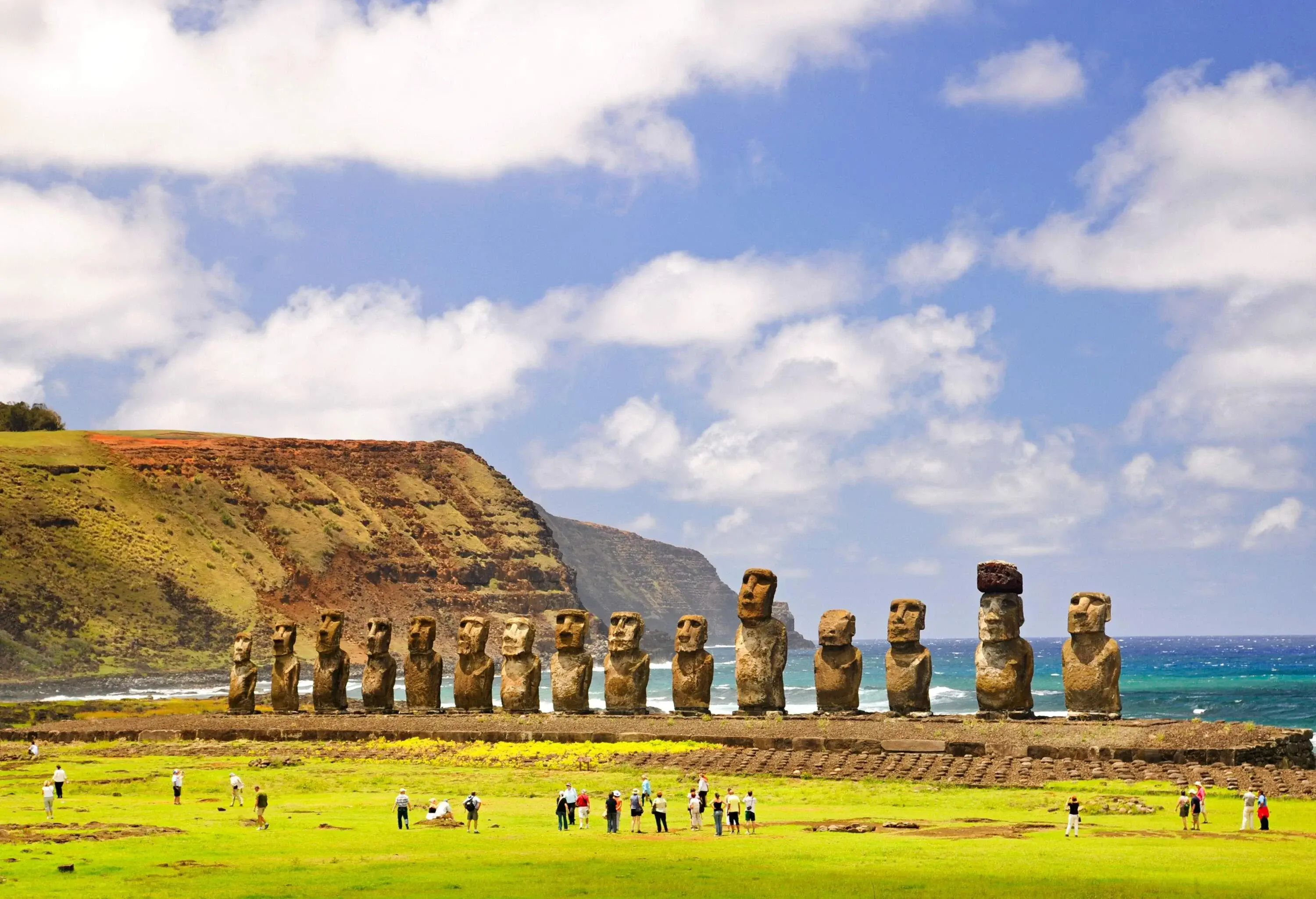 The iconic 15 Moai statues, large stone monoliths carved by the Rapa Nui people, stand tall along the coastline, with visitors exploring the nearby turf and a dramatic coastal cliff serving as the backdrop.