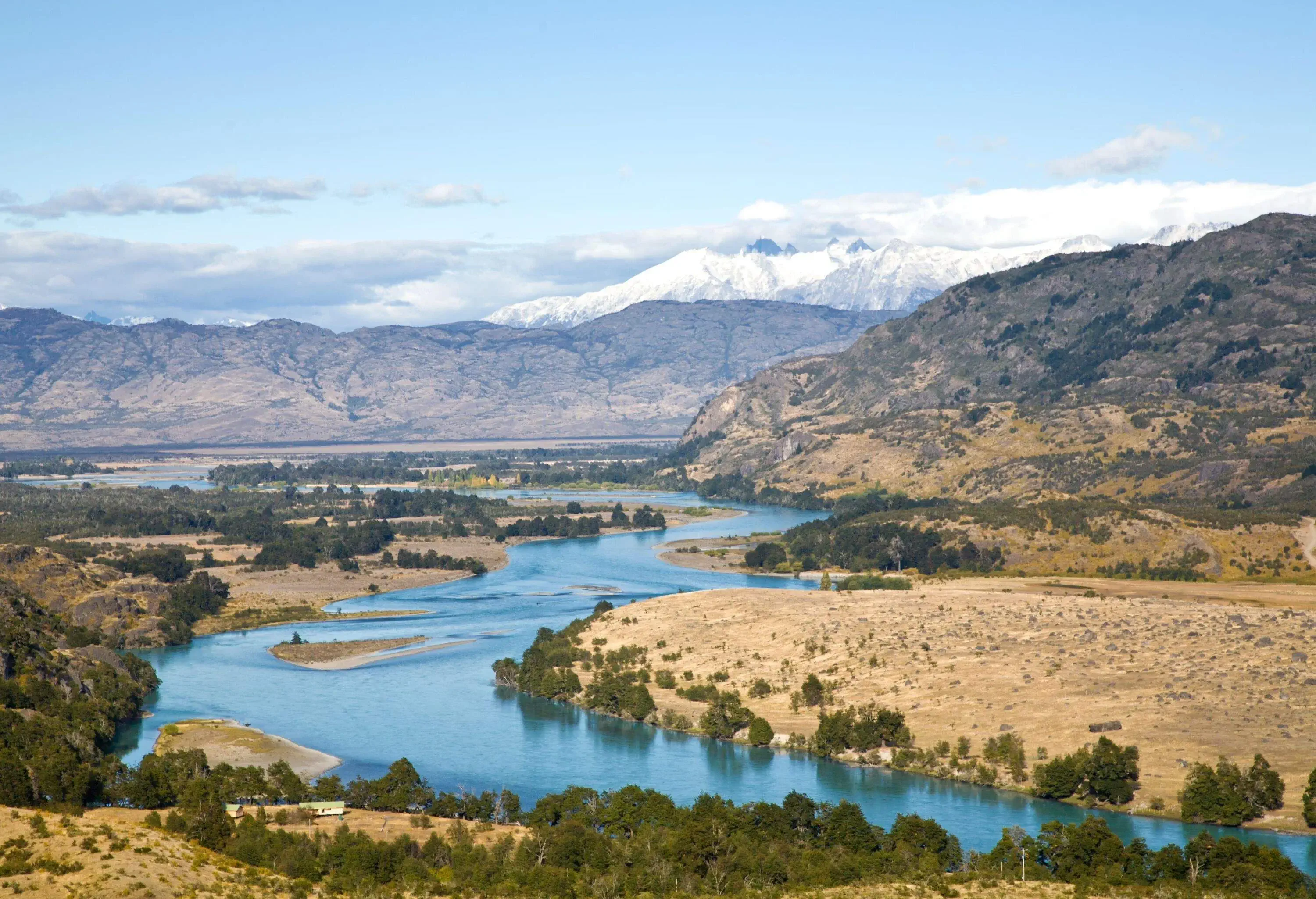 Panoramic view of river bed surrounded by greenery in a mountainous valley, with snow sprinkled peak in the background