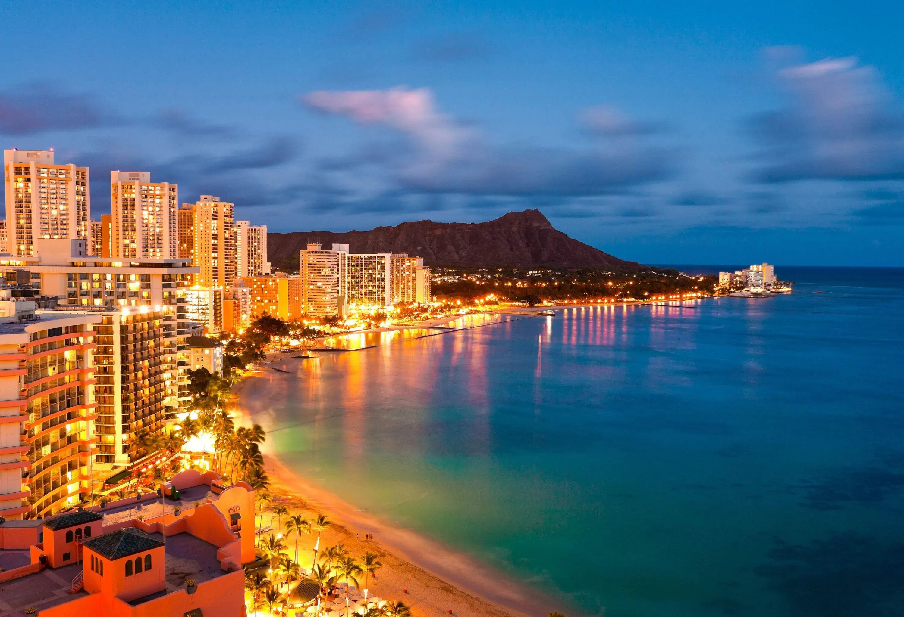 The coast comes alive with towering buildings standing tall and brightly illuminated, while the iconic Diamond Head looms in the background.