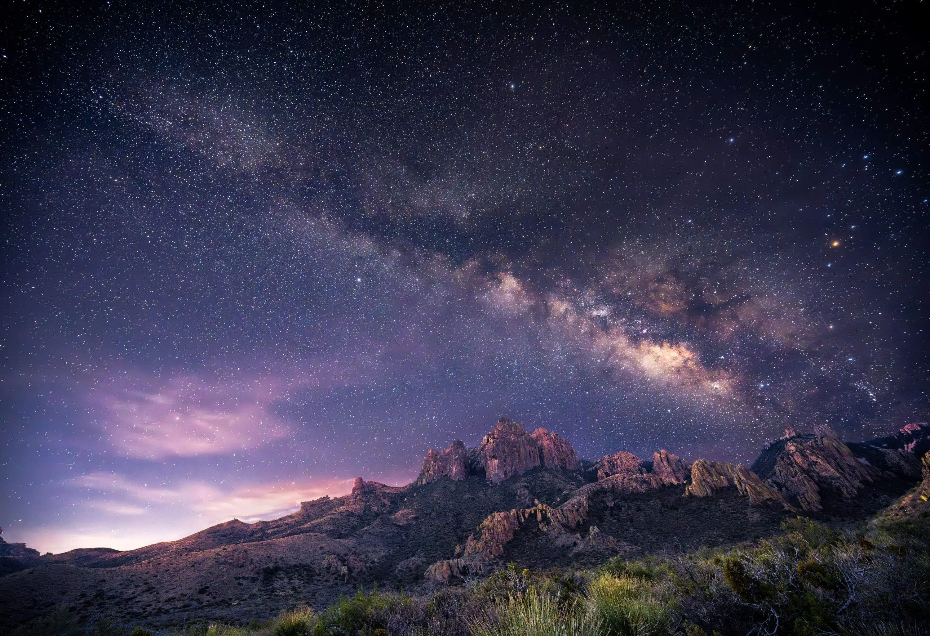 a stunning night sky with the Milky Way galaxy arching above rugged, illuminated mountains. The scene has a purplish hue, highlighting the stars and the dramatic landscape.
