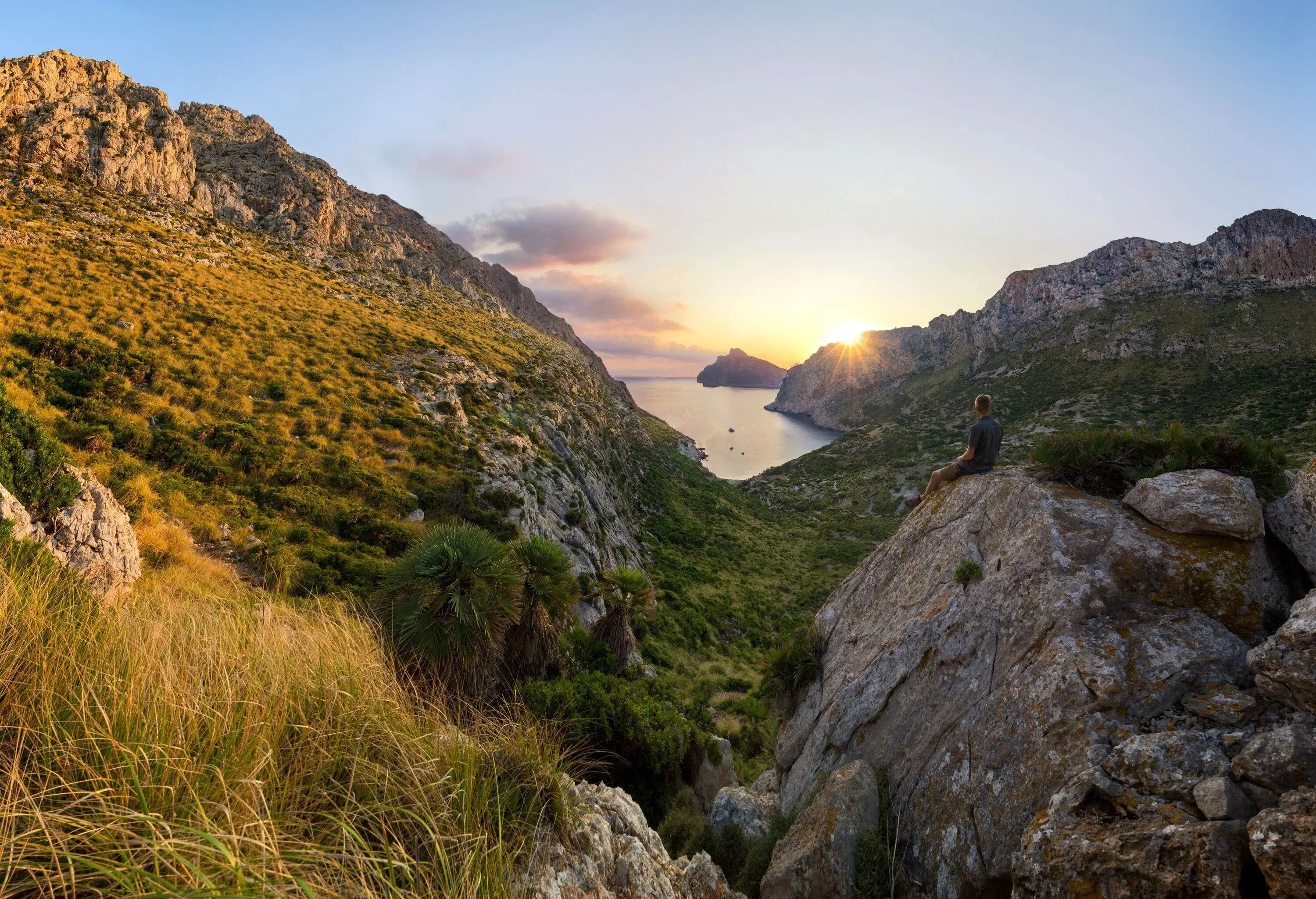 Man watches the sunrise through the mountains as he sits on a boulder near a grassy mountain slope.