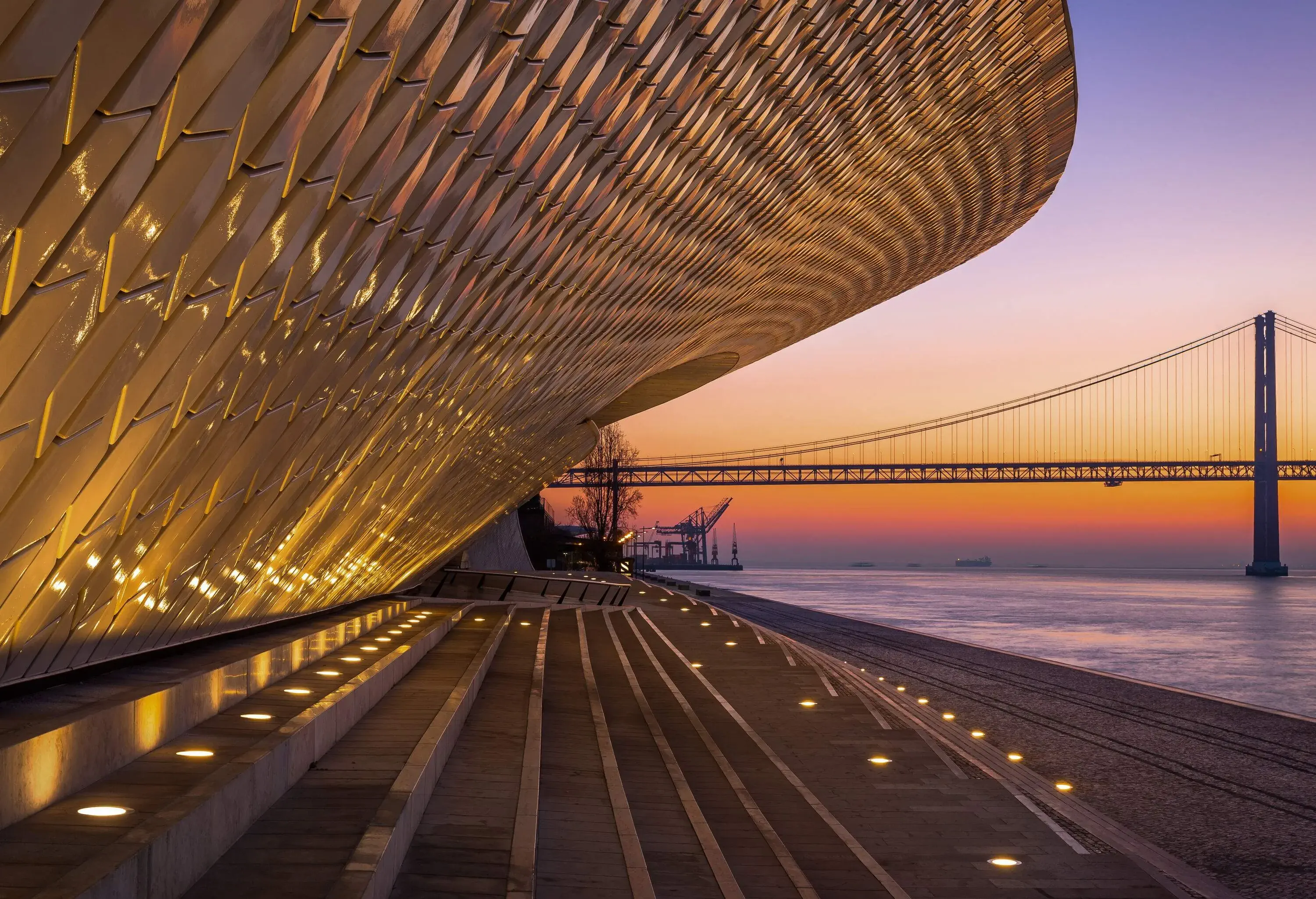 An illuminated unique outward-looking museum on the riverbank with a view of a long suspension bridge at sunset.