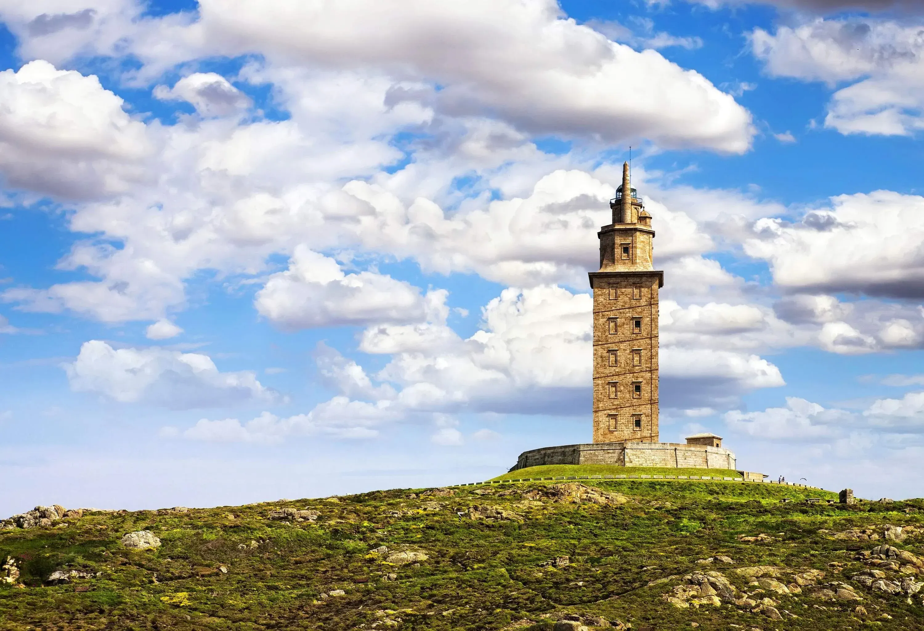 The Tower of Hercules is a tall lighthouse with a spiral ramp outline situated on a grassy hill.