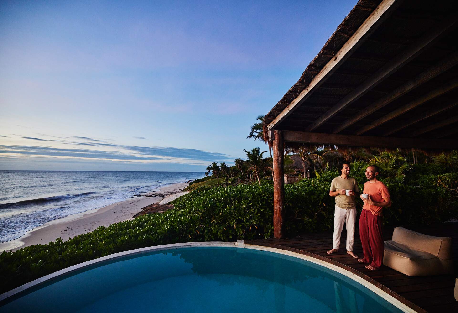 Wide shot of gay couple standing poolside watching sunrise at luxury tropical villa overlooking beach and ocean, Mexico