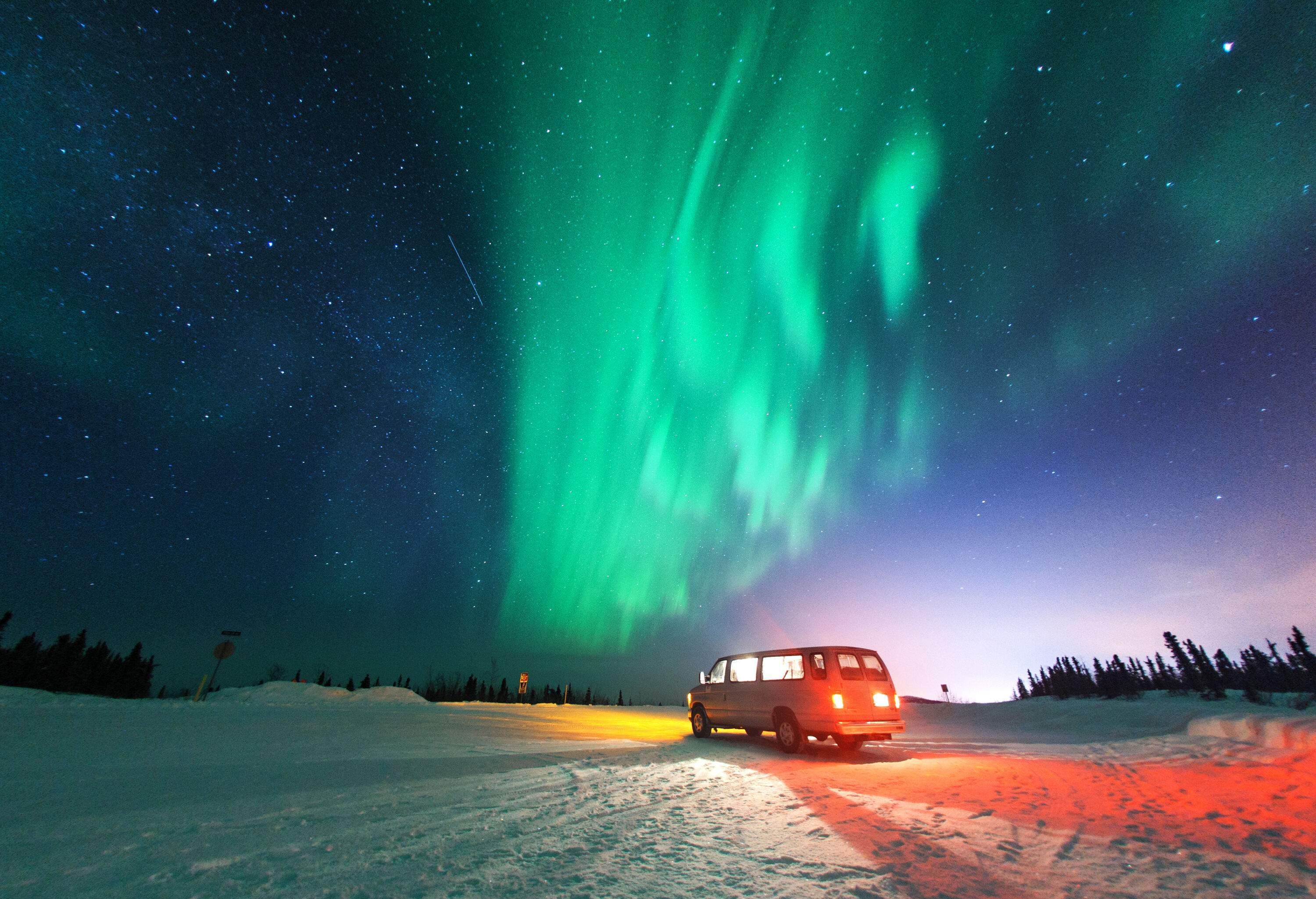 A van parked in a snowfield under the blazing northern lights and twinkling stars.