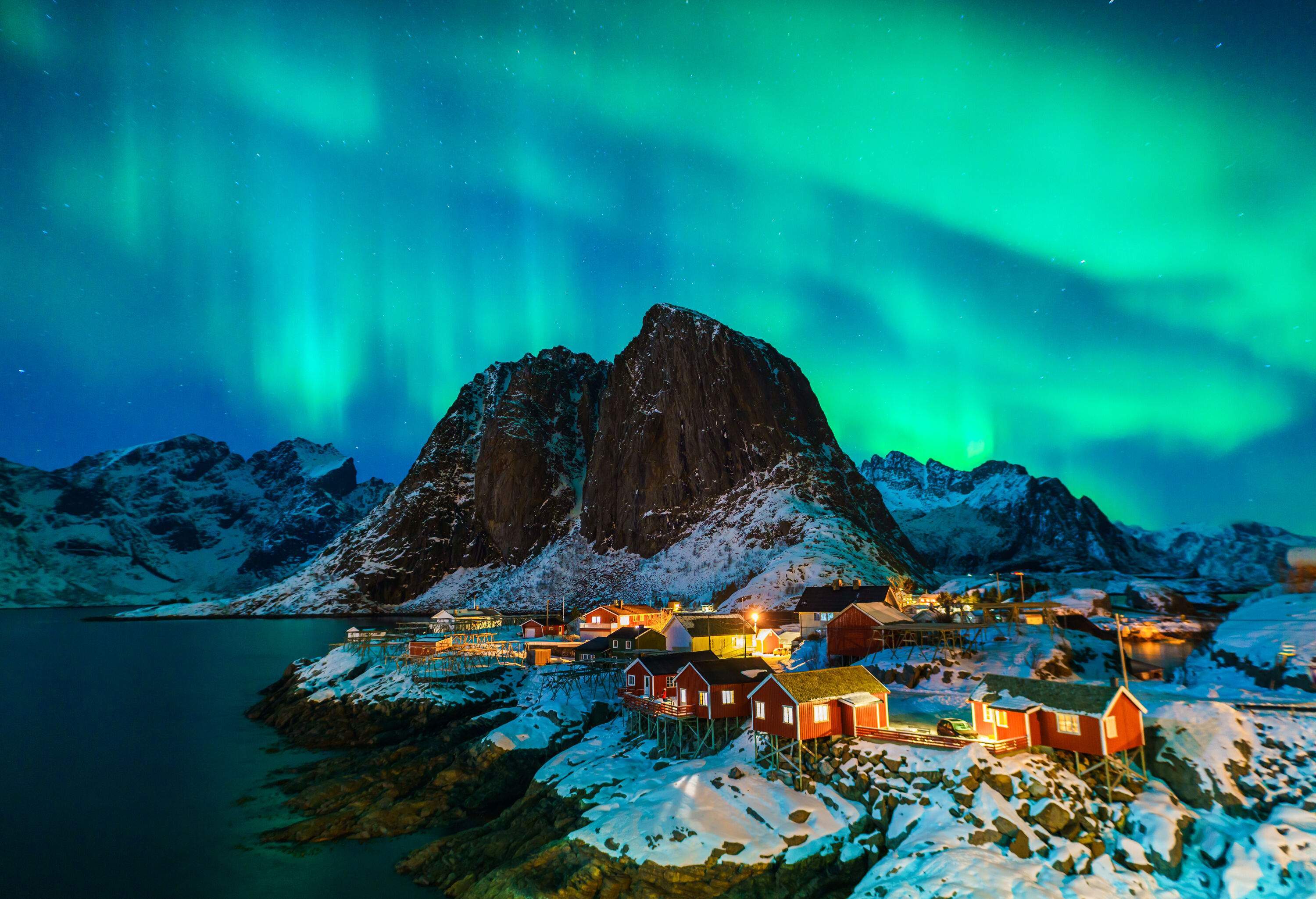 Green northern lights dancing across the sky over a fishing village with cabins across the rocky and snow-covered coast.