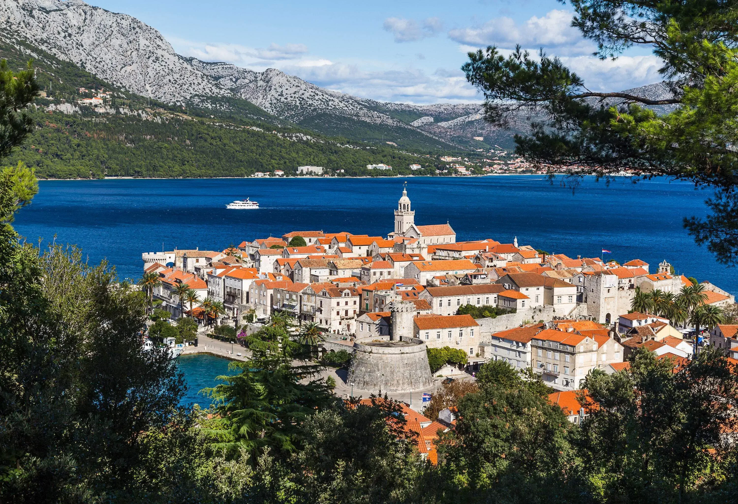 A picturesque old town with classic stone houses and buildings jutting out into the turquoise sea overlooking a lush mountain range.