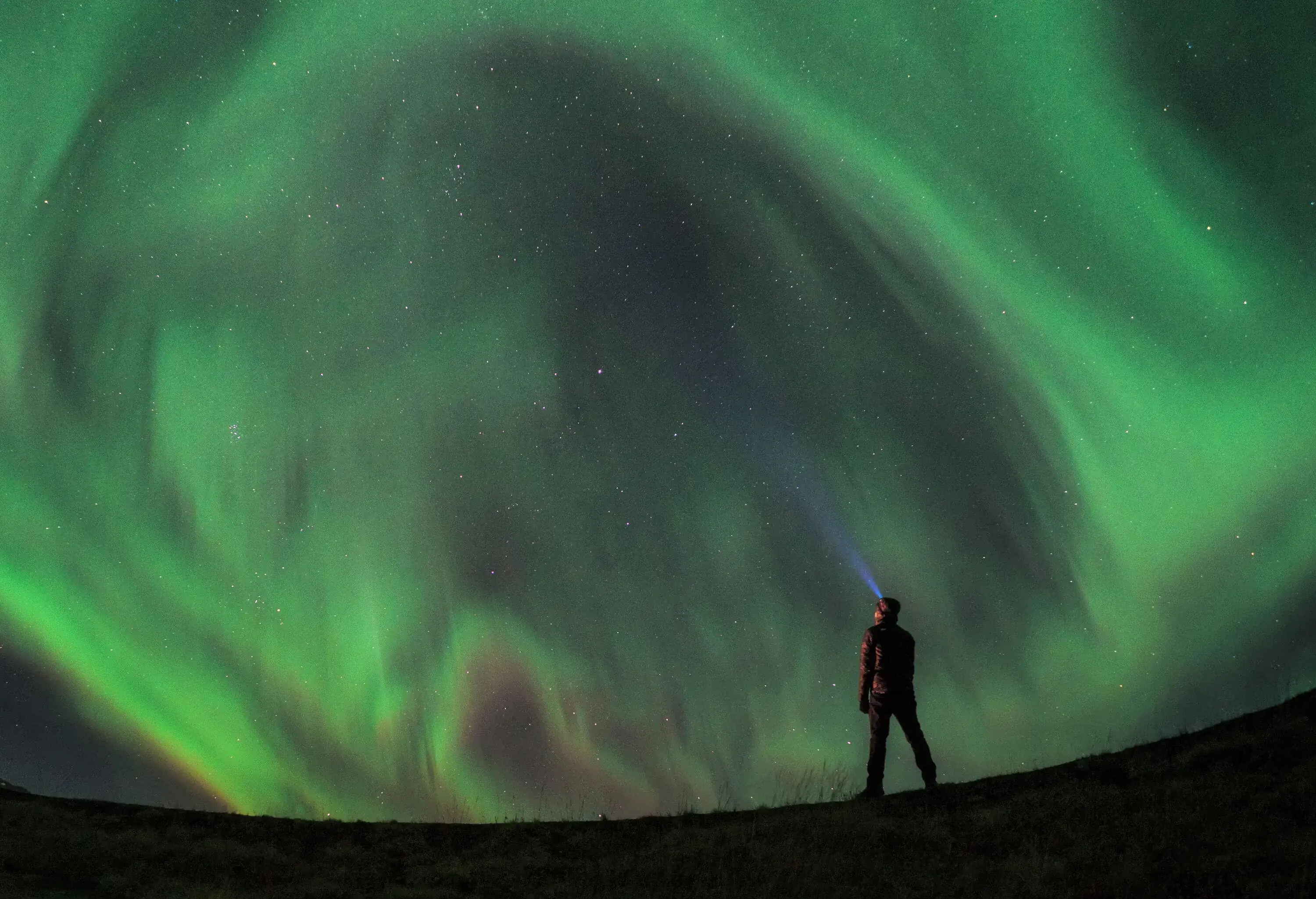 A man with a lit headlamp stands in the foreground, amazed by the Northern lights dazzling in the sky.
