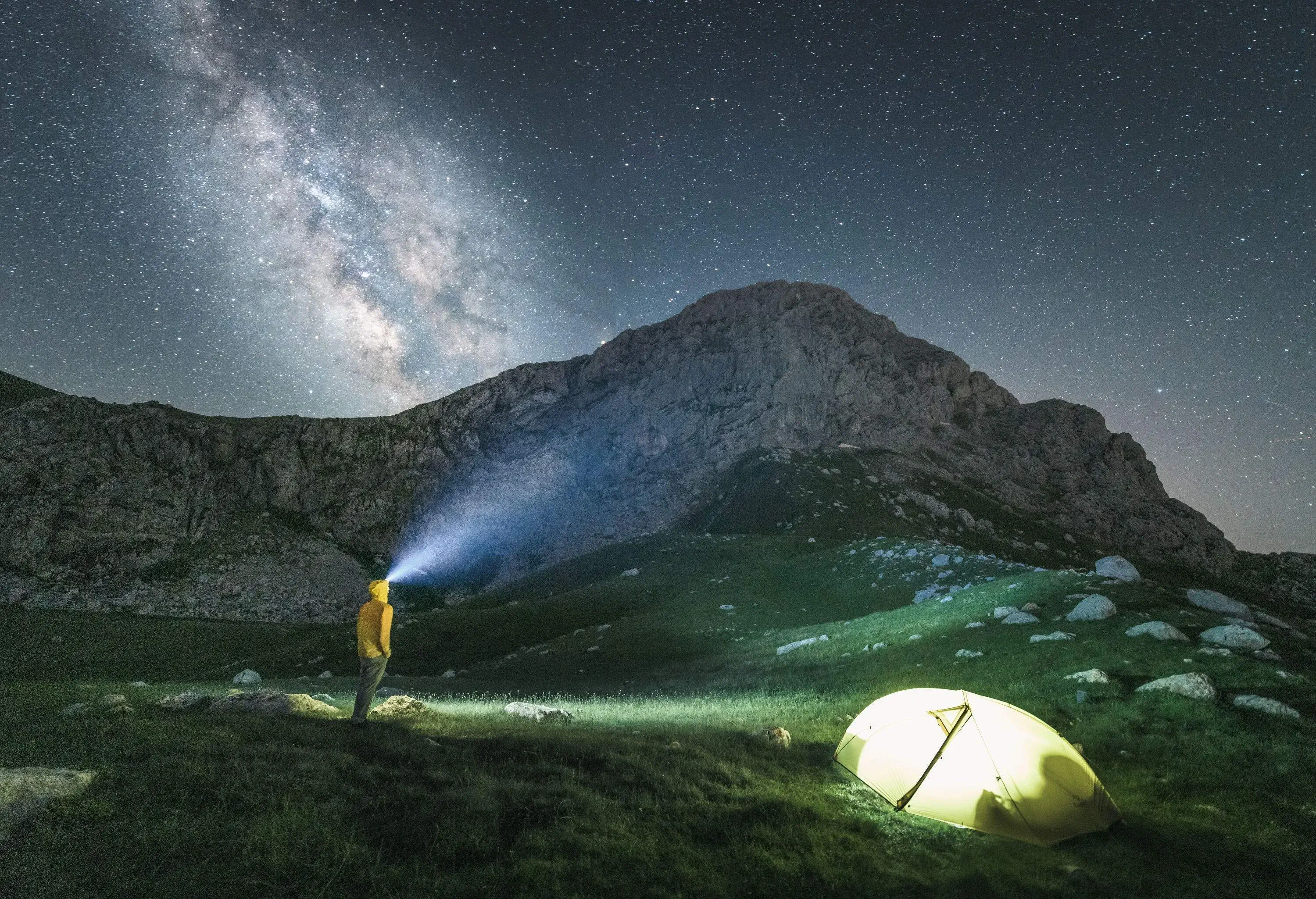 A night-time view of a man with a headlight standing next to a bright tent under a constellation of stars.