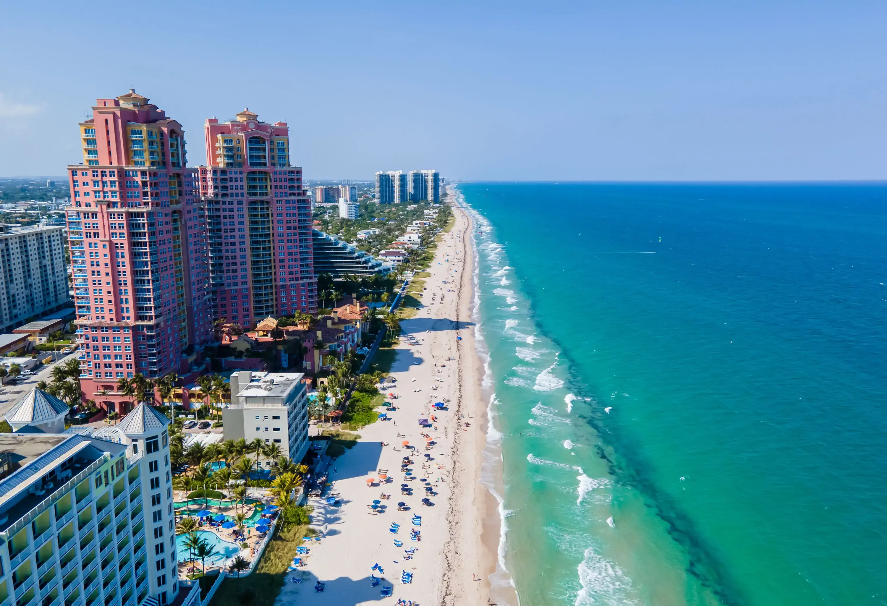 A crowded white sand beach with turquoise waters along a cluster of hotels, resorts, and residential buildings.
