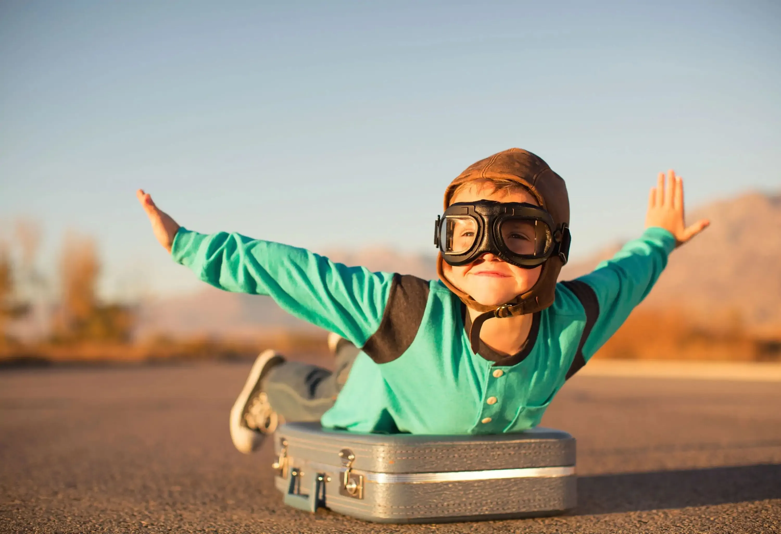 A young boy wearing a flight cap and goggles lies on top of a suitcase with outstretched arms, joyfully imagining he is flying, his face adorned with a wide smile.