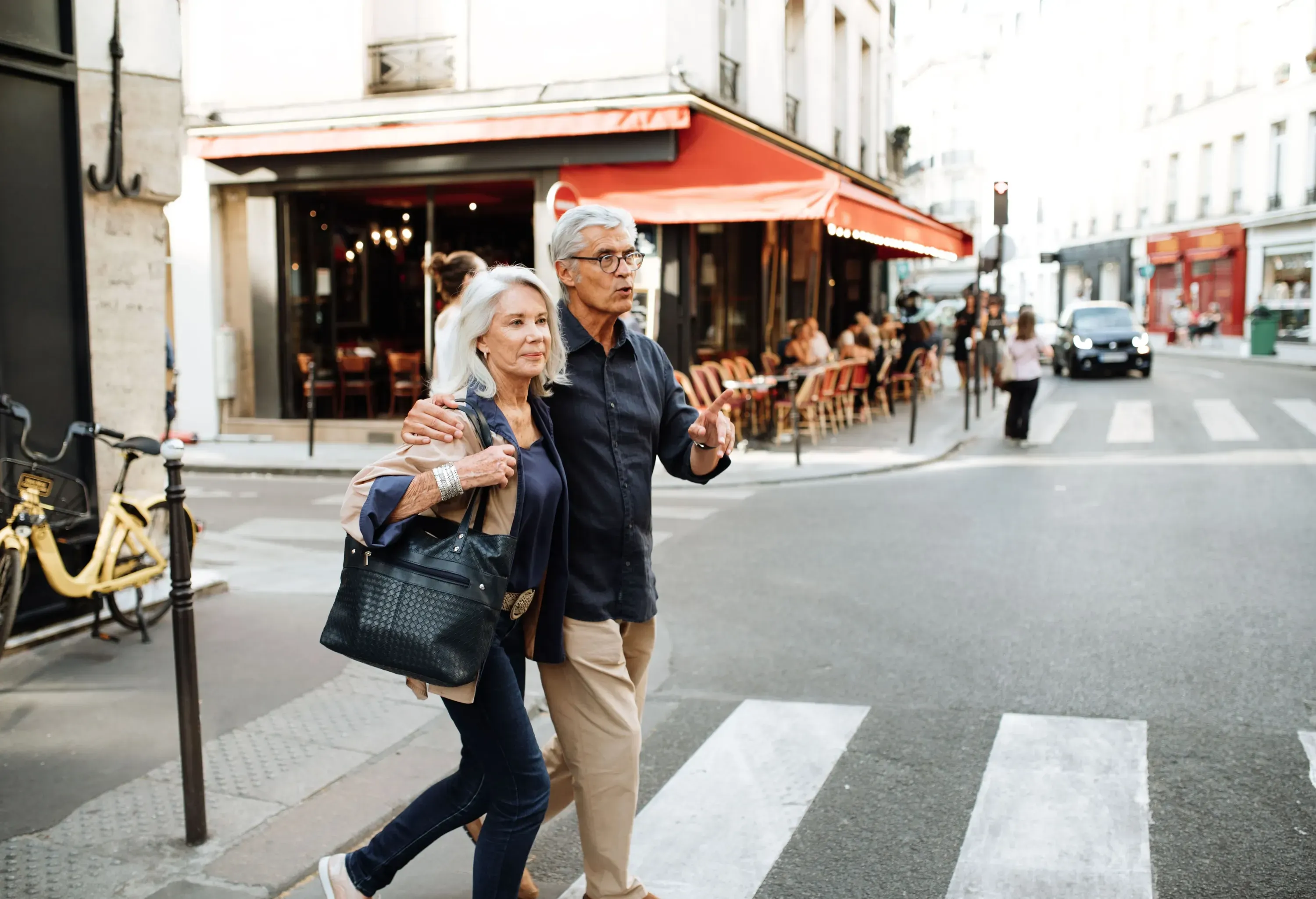 A lovely senior couple gracefully crosses the pedestrian lane, with a charming café building standing proudly in the background.