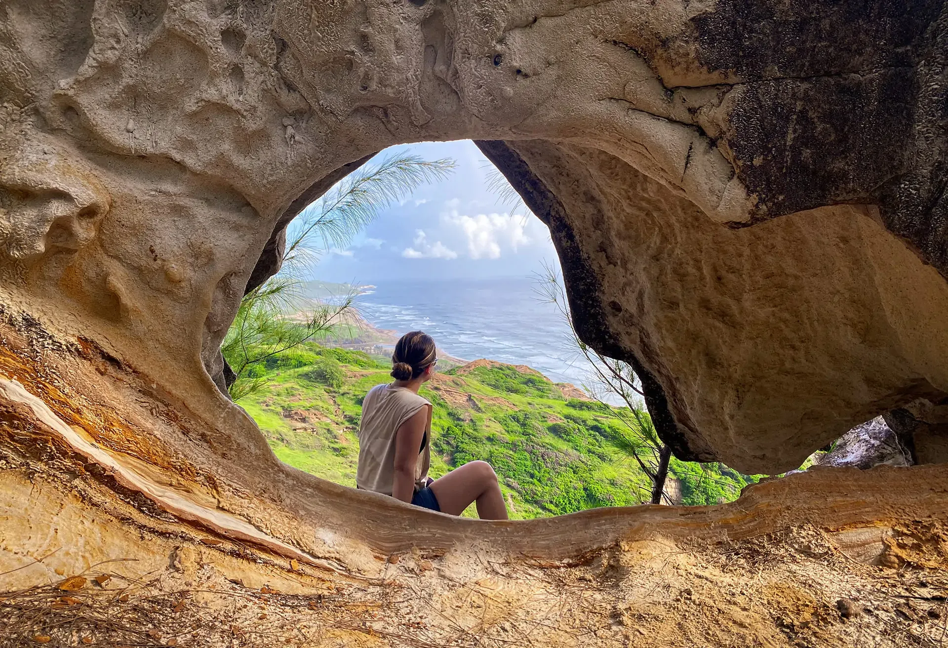 A woman sitting inside a rock cave opening, looking out at a lush green landscape and the ocean.
