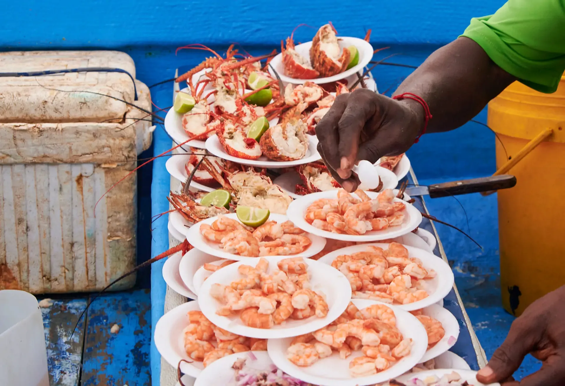 A vendor serving fresh shrimp and lobster on plates on a boat with lime wedges.