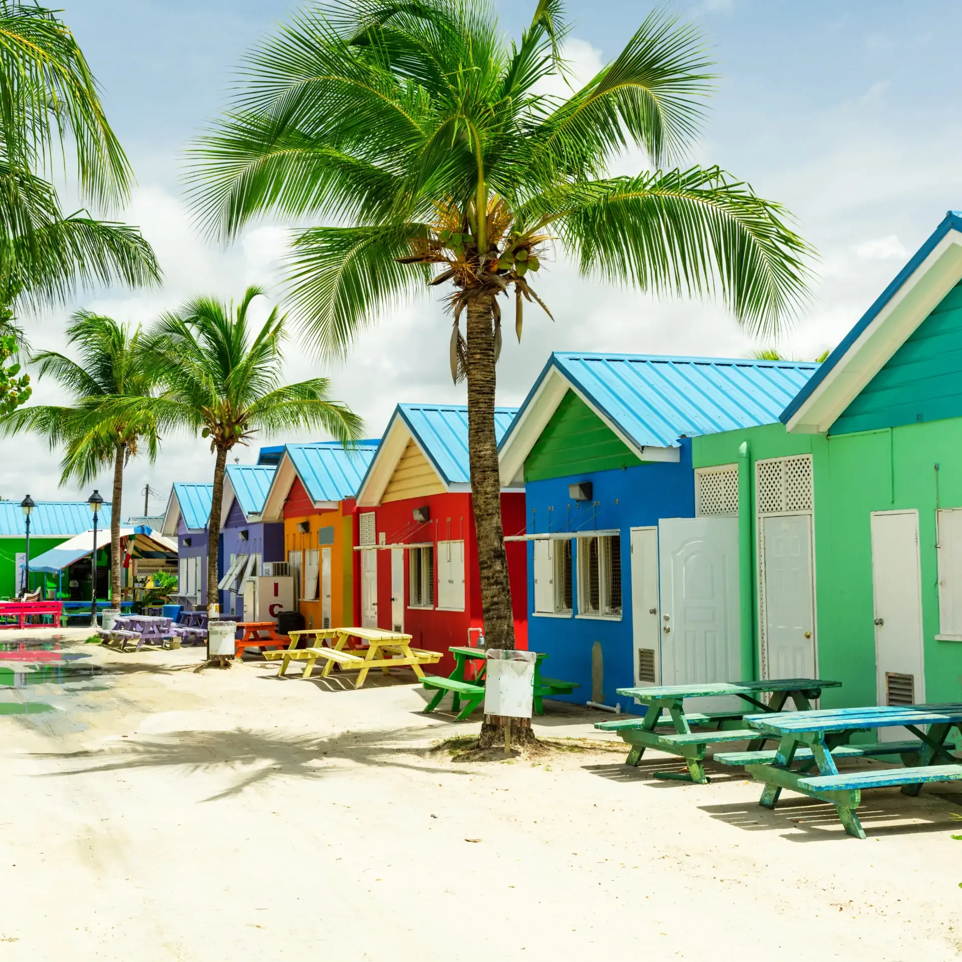 Brightly colored beach huts and palm trees on white sand under a clear sky.