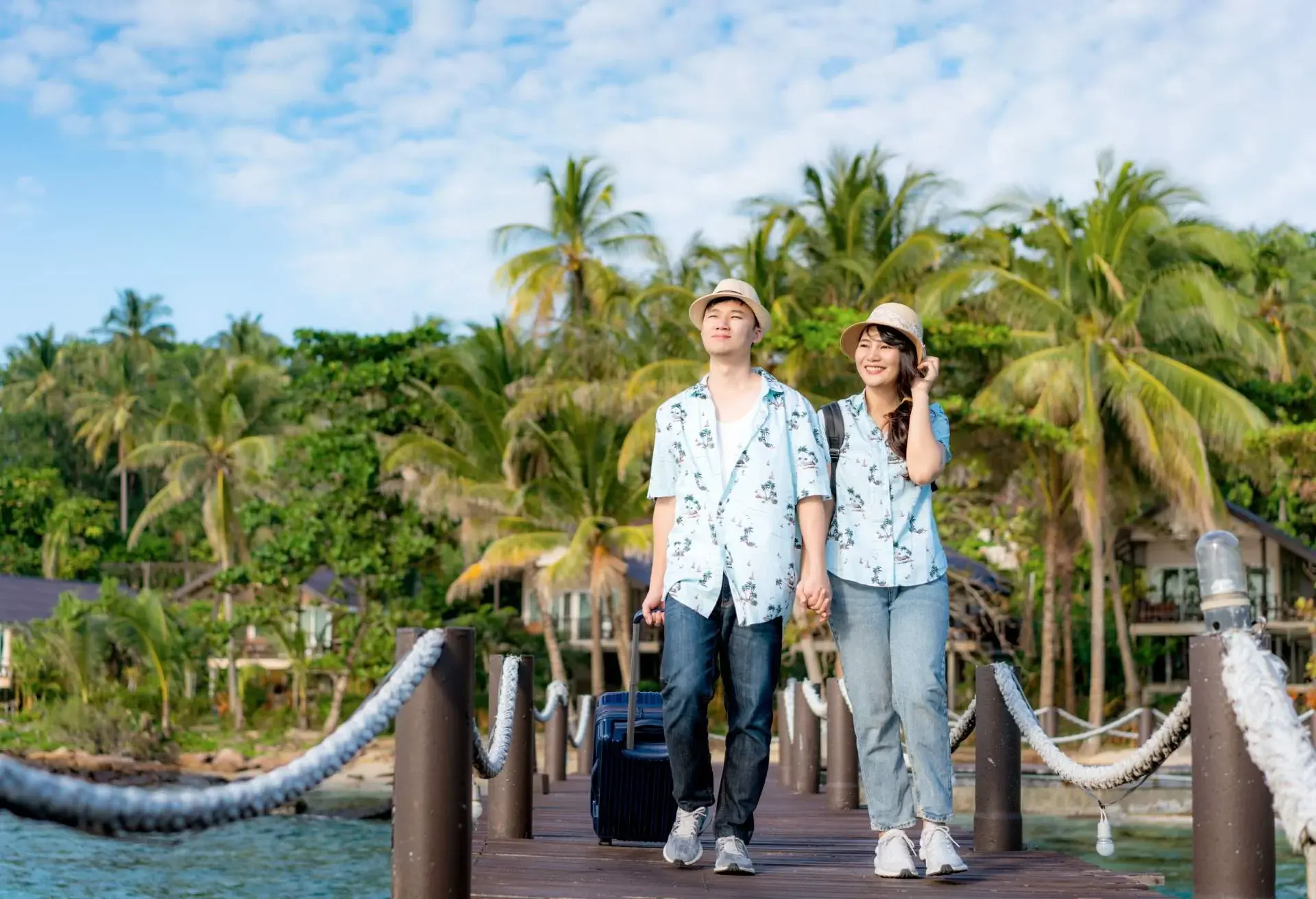 A man and woman walk down a dock with suitcases. Palm trees and a blue sky are in the background.