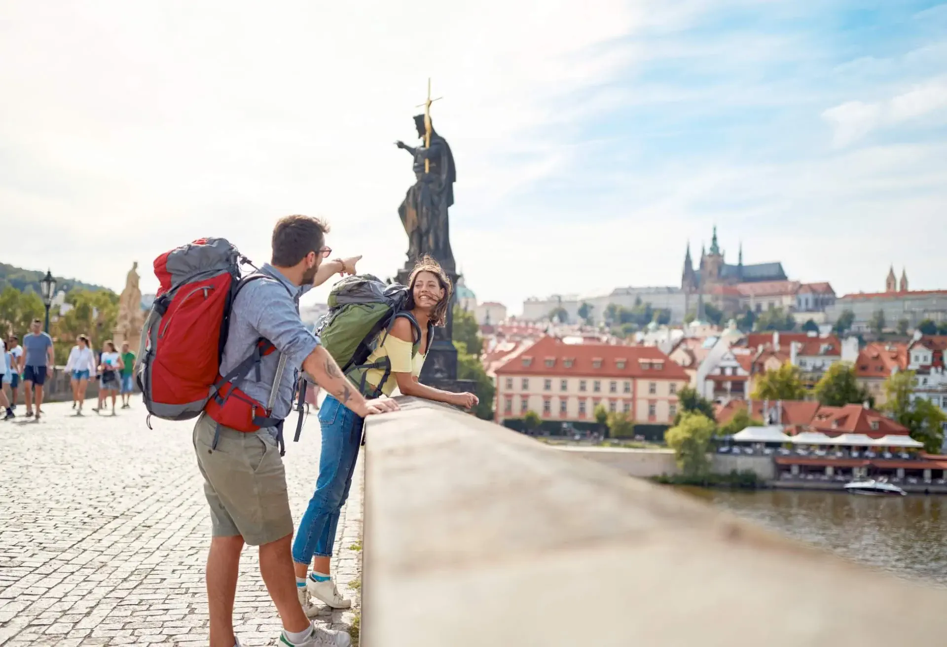 A young man and woman lean over a bridge in Europe. The man points at the view in the distance while the woman looks at him, smiling.