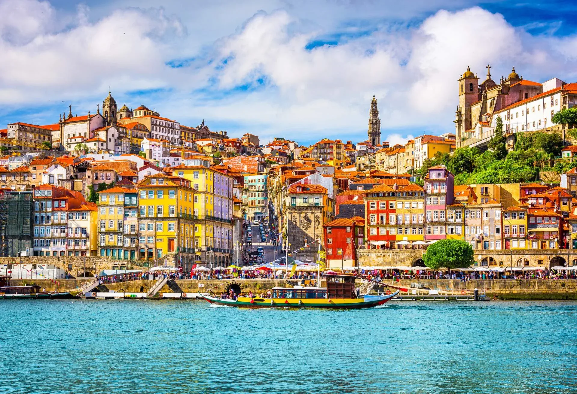 A cluster of colourful and vibrant buildings in a riverside city along a bustling promenade and harbour with cruising boats.