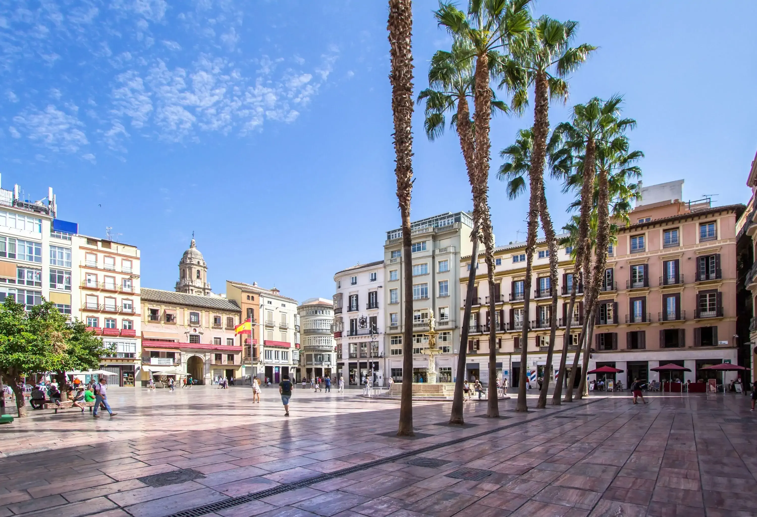 People strolling through a public square with a fountain and a flag flying on a pole.