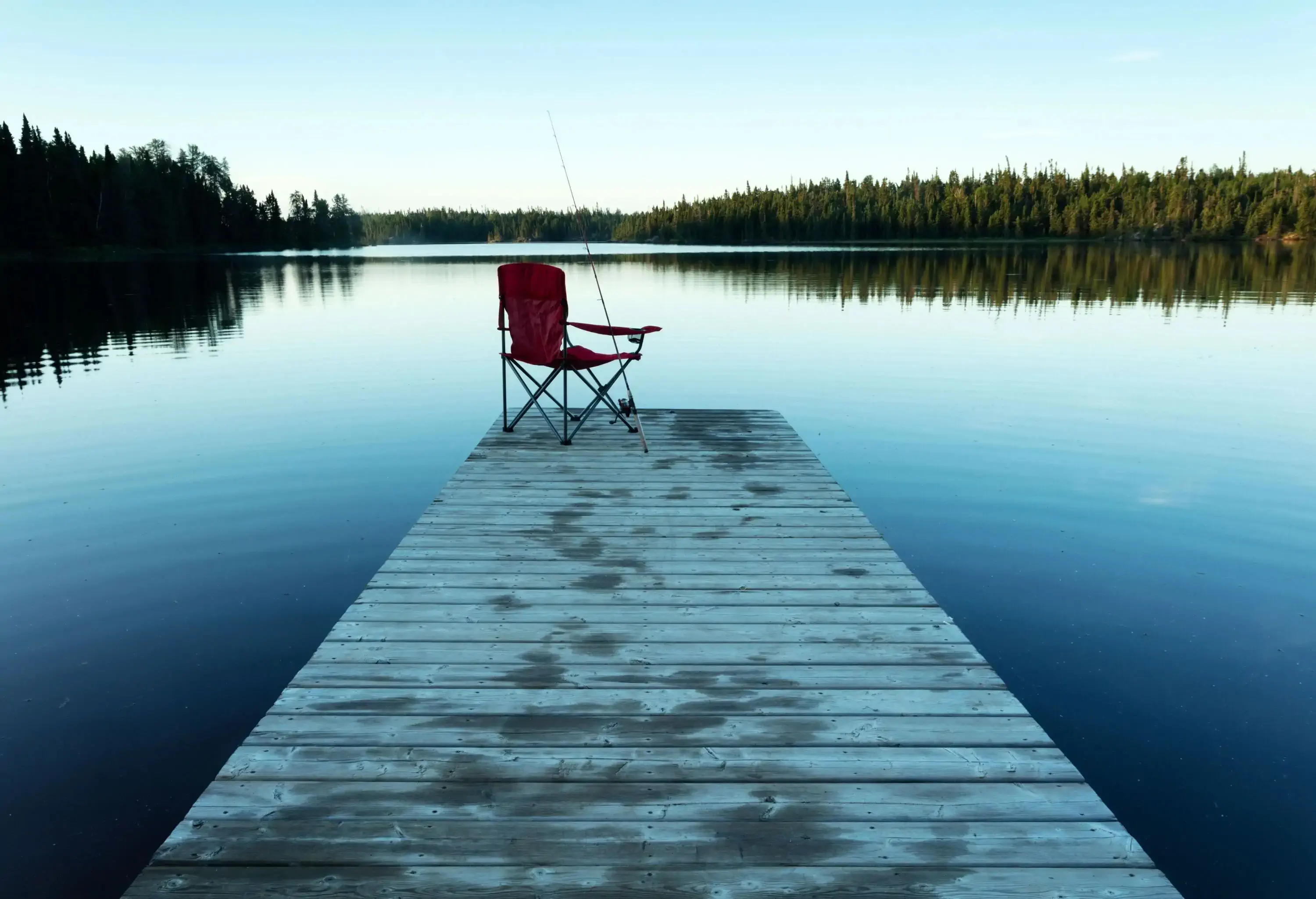 A chair with a fishing rod set at the edge of a jetty in front of a calm lake.
