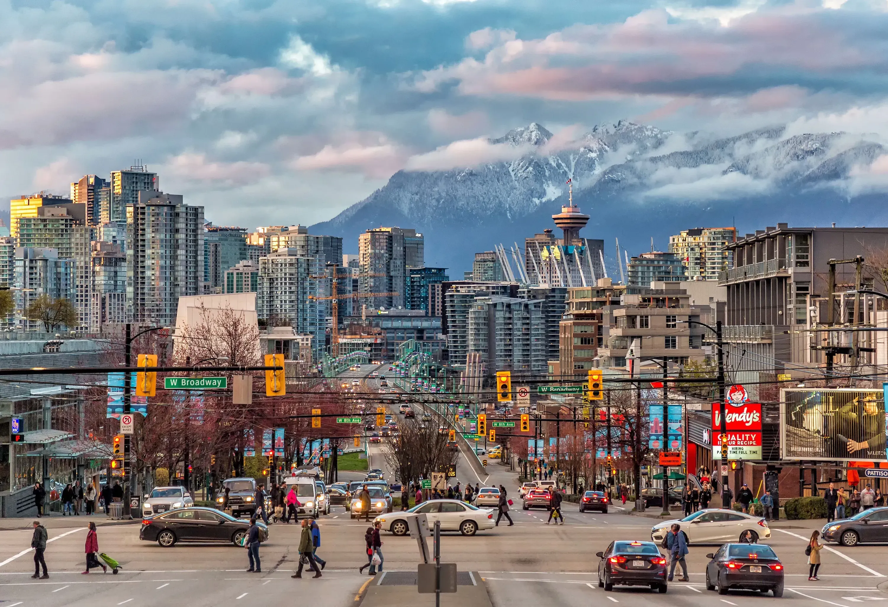 A bustling city centre skyline with people and cars moving through the streets with snowy mountains in the background.