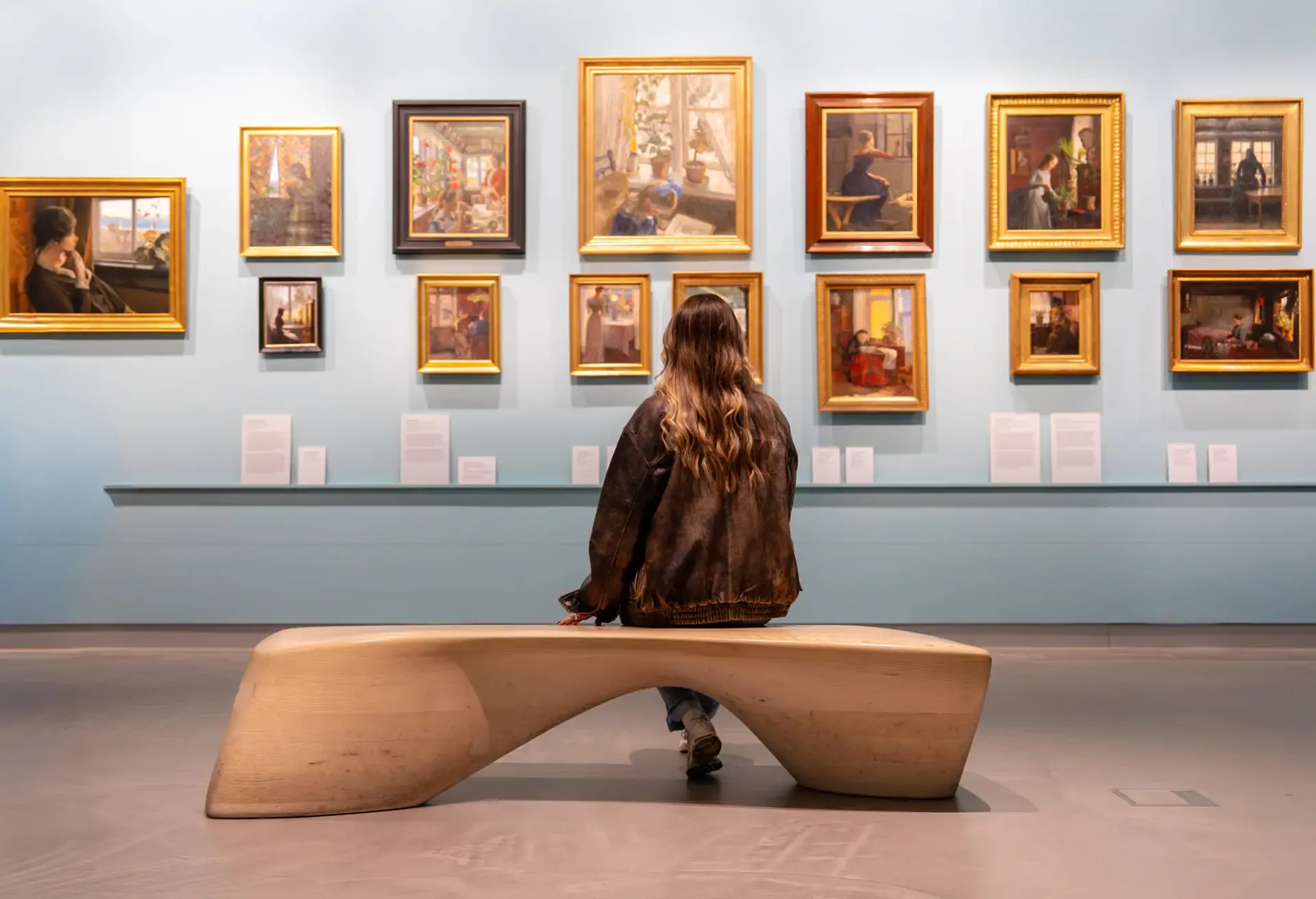 Woman sitting on a bench in an art gallery looking at framed paintings.