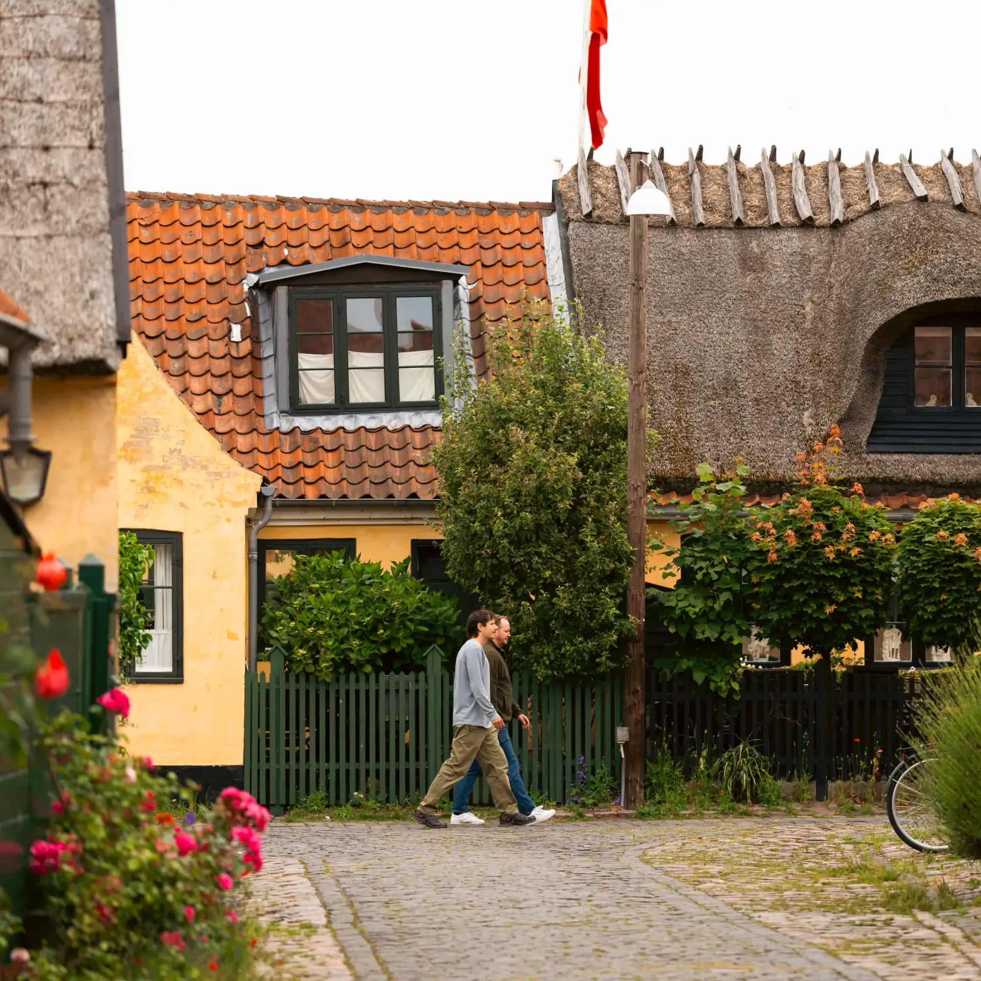 Two men walking down a cobblestone street lined with traditional yellow houses and thatched roofs.
