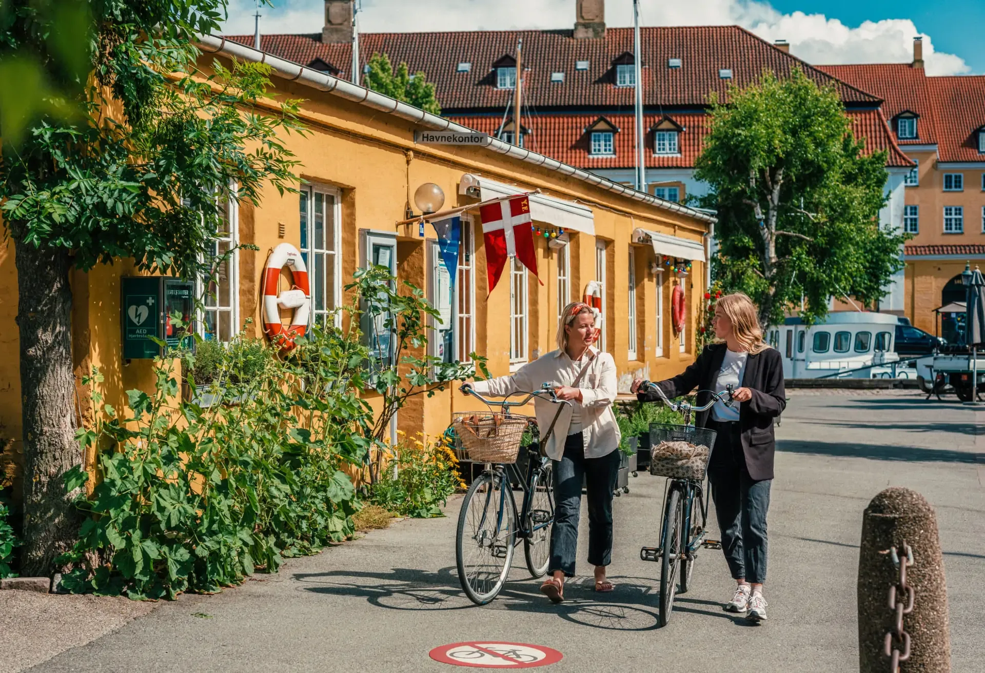 Two women walking bikes past a yellow building with a Danish flag.