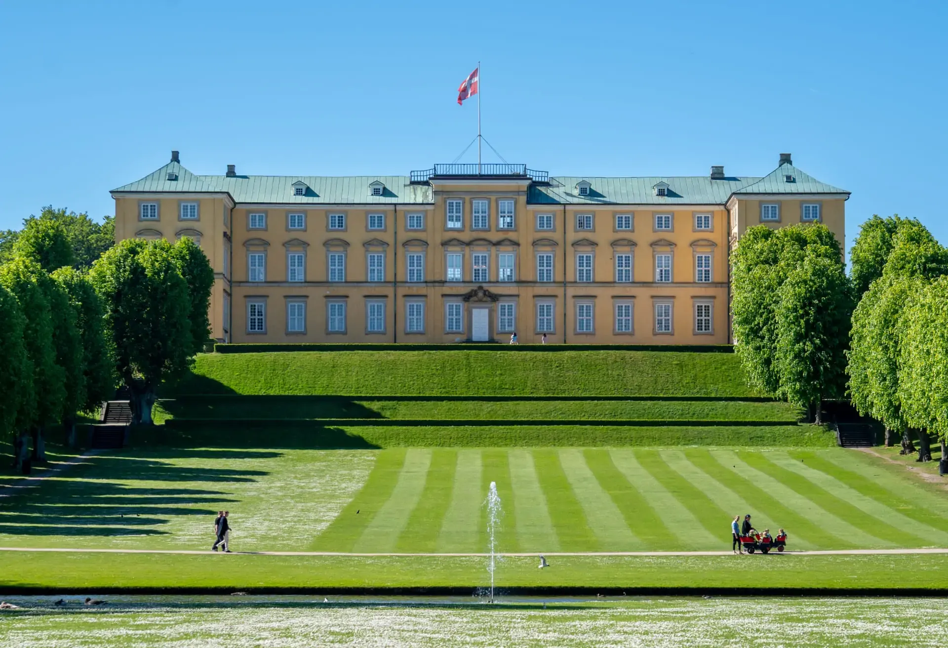 Yellow palace on a hill with terraced lawns and a fountain in the foreground.