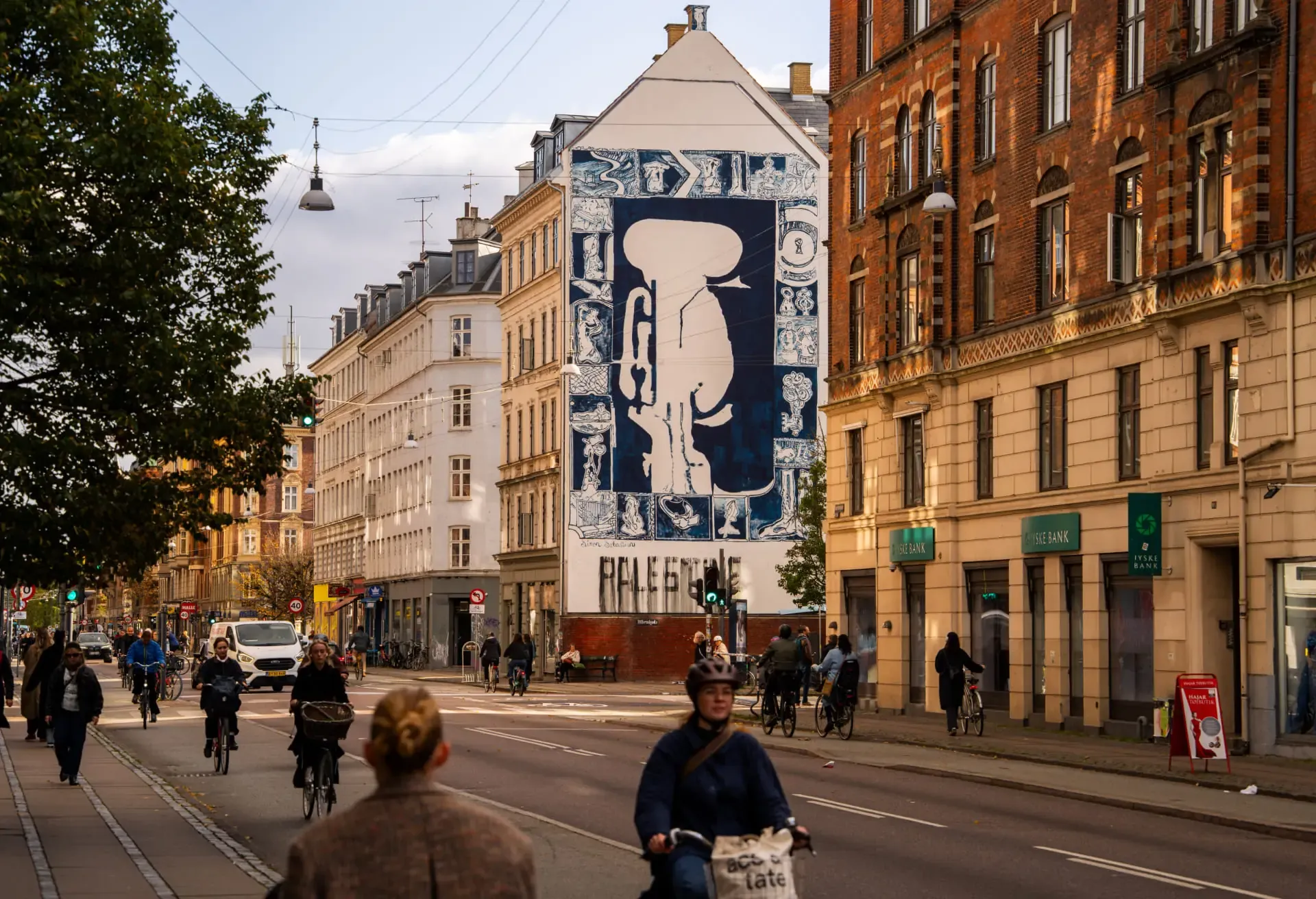 Cyclists riding on a city street past a large blue and white mural on a building.