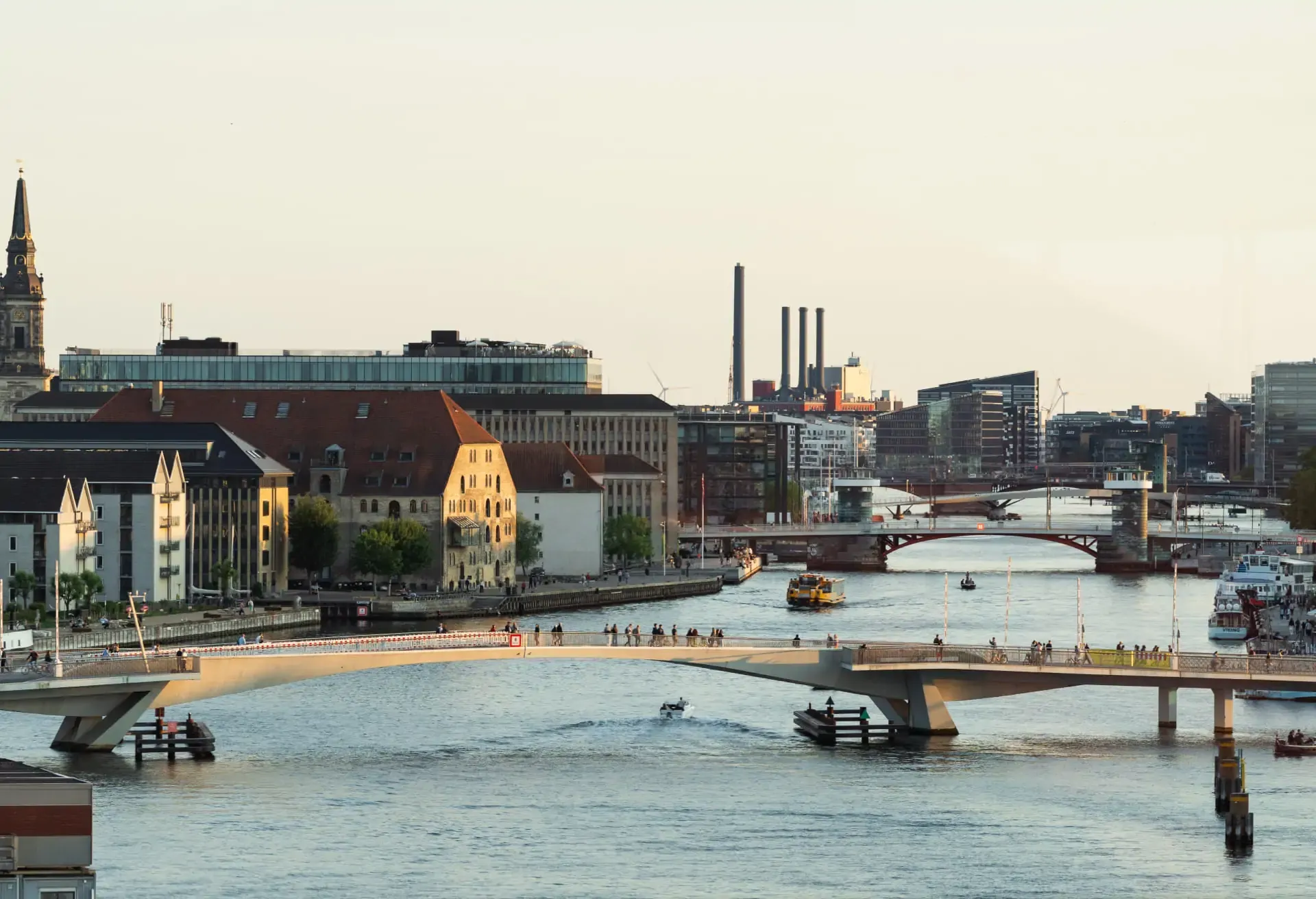 Modern bridge over a city canal with people, boats, and historic architecture.