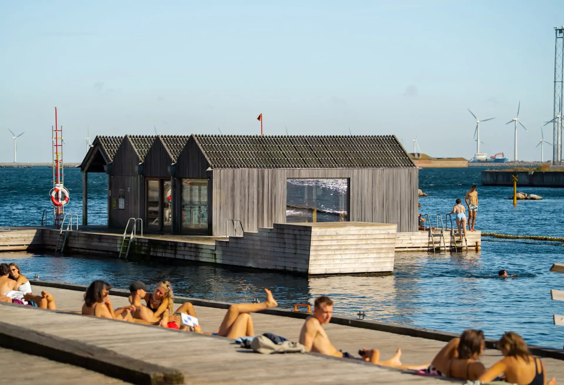 People sunbathing at a harbor bath with wind turbines in the background.