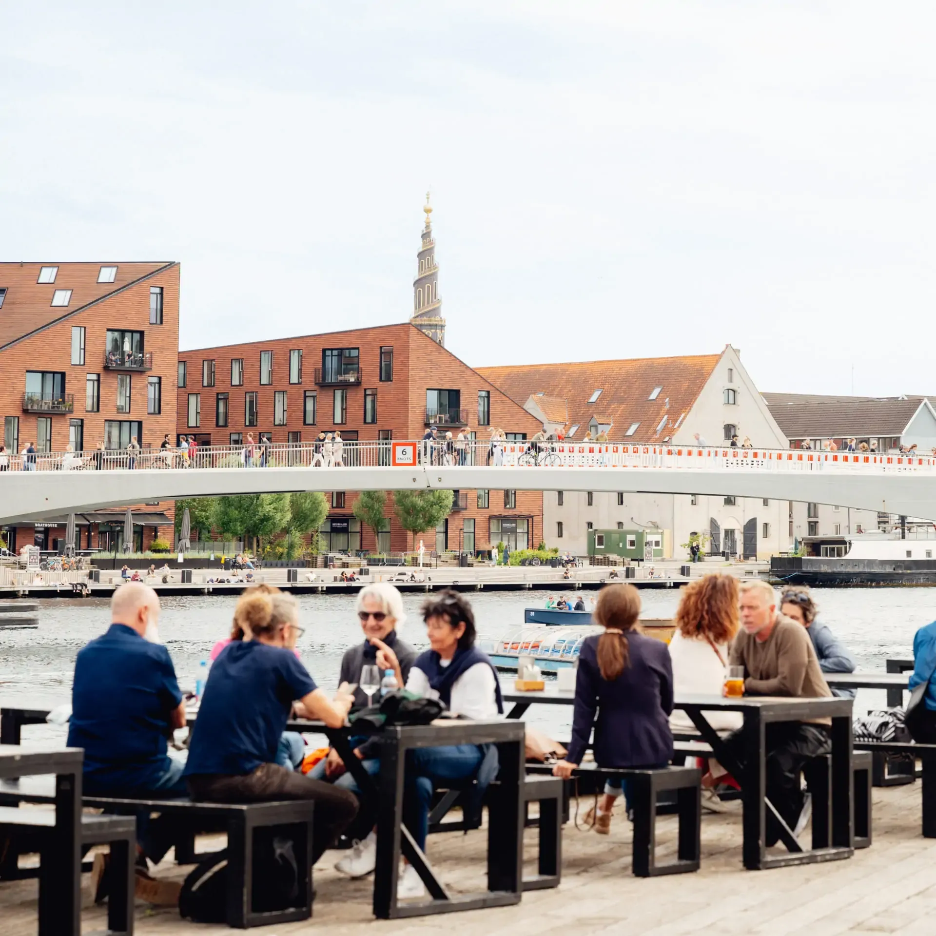 People relaxing at outdoor tables by a city canal with a modern bridge and historic tower.